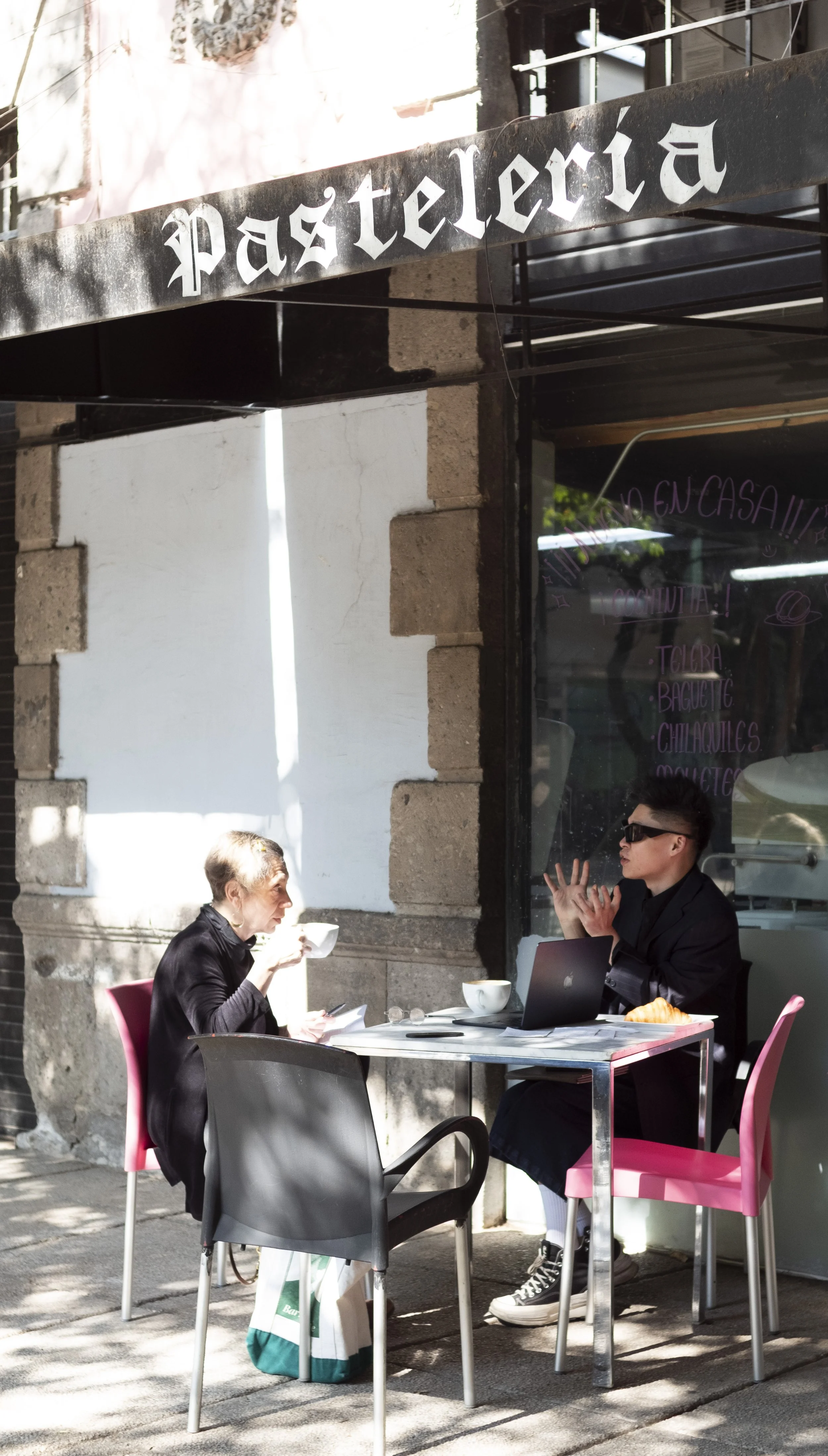 Two people dining outside a cafe called "Pasteleria." One woman with short gray hair is eating from a bowl, and a person with sunglasses and dark hair is talking and gesturing while seated at the table. There are pink and black chairs, a laptop, and 