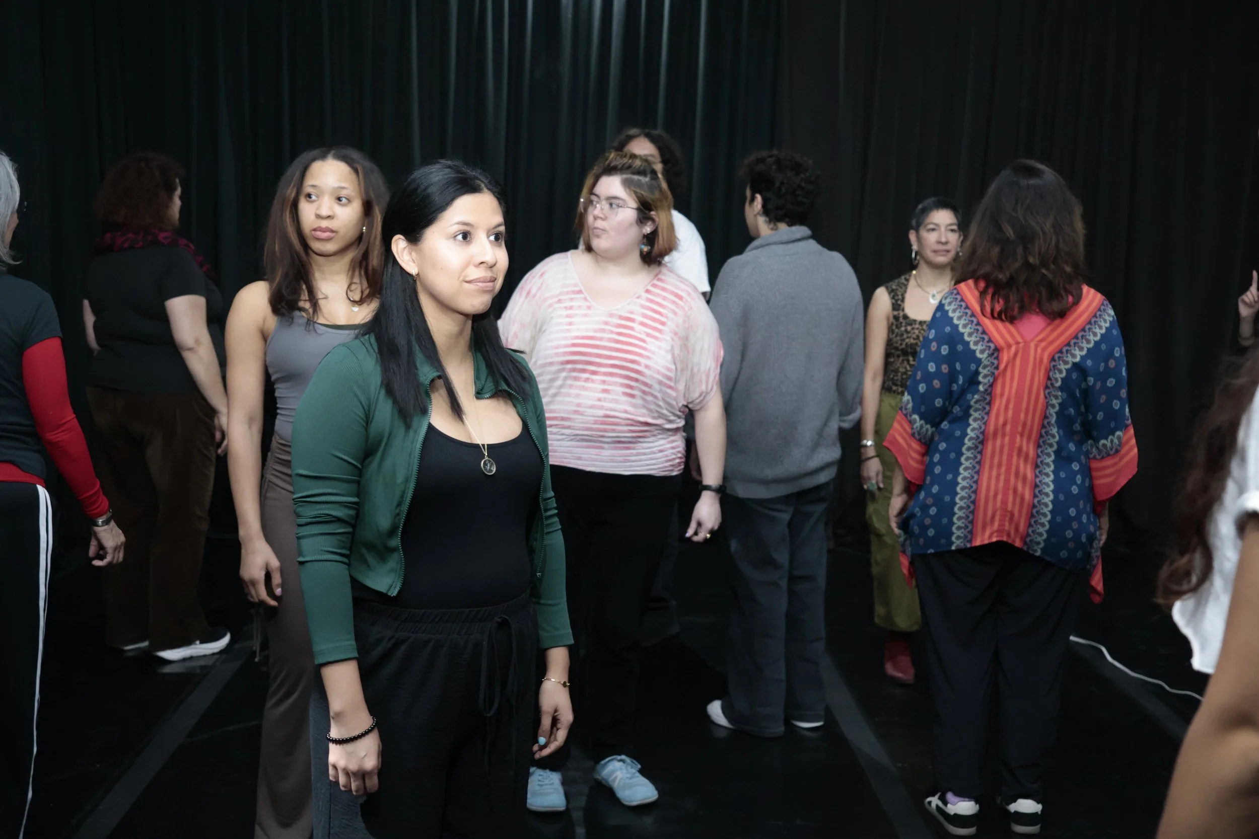 A group of diverse women standing and talking in a room with dark curtains in the background.