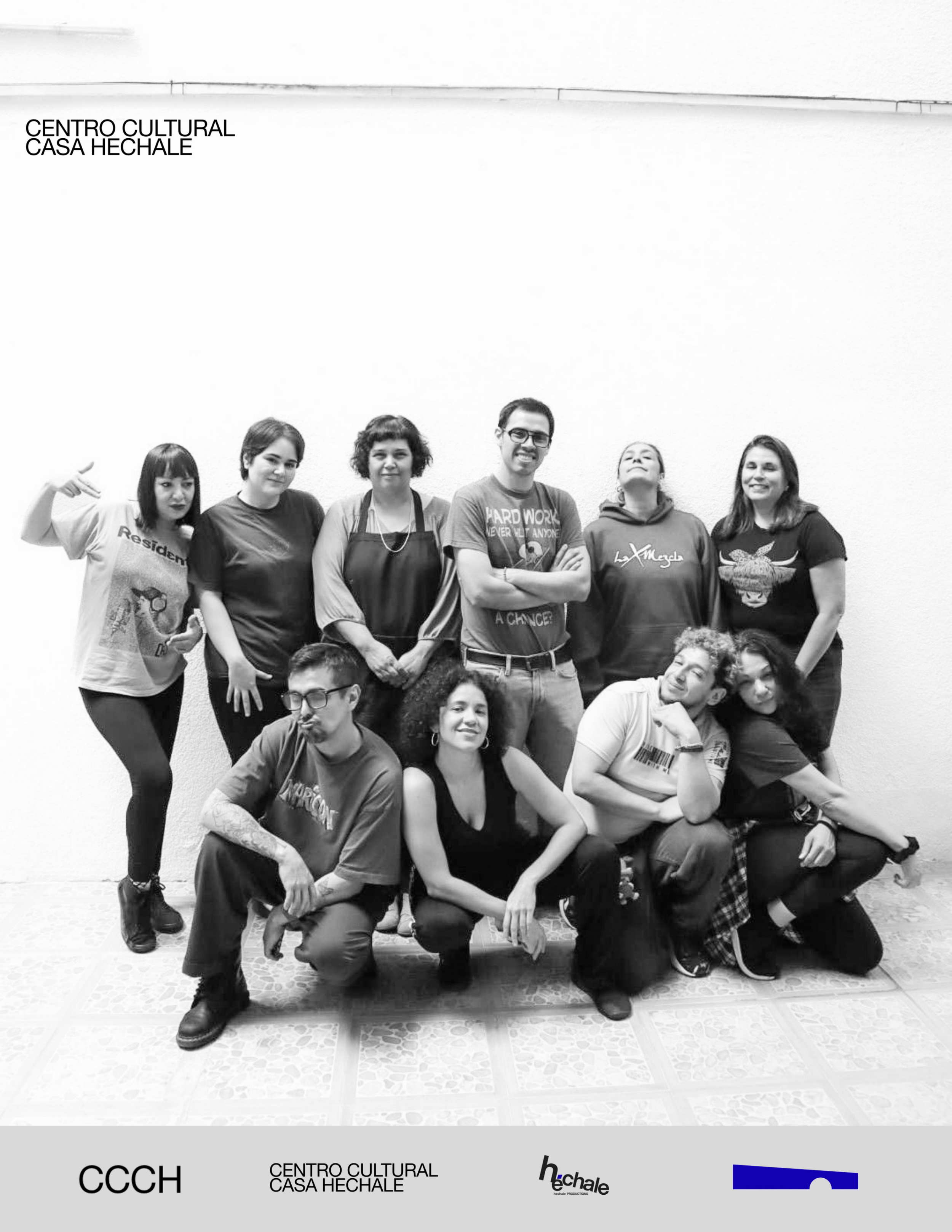 A black and white group photo of seven people standing and sitting in front of a plain wall. Some individuals are smiling or striking playful poses, and one person is wearing glasses.