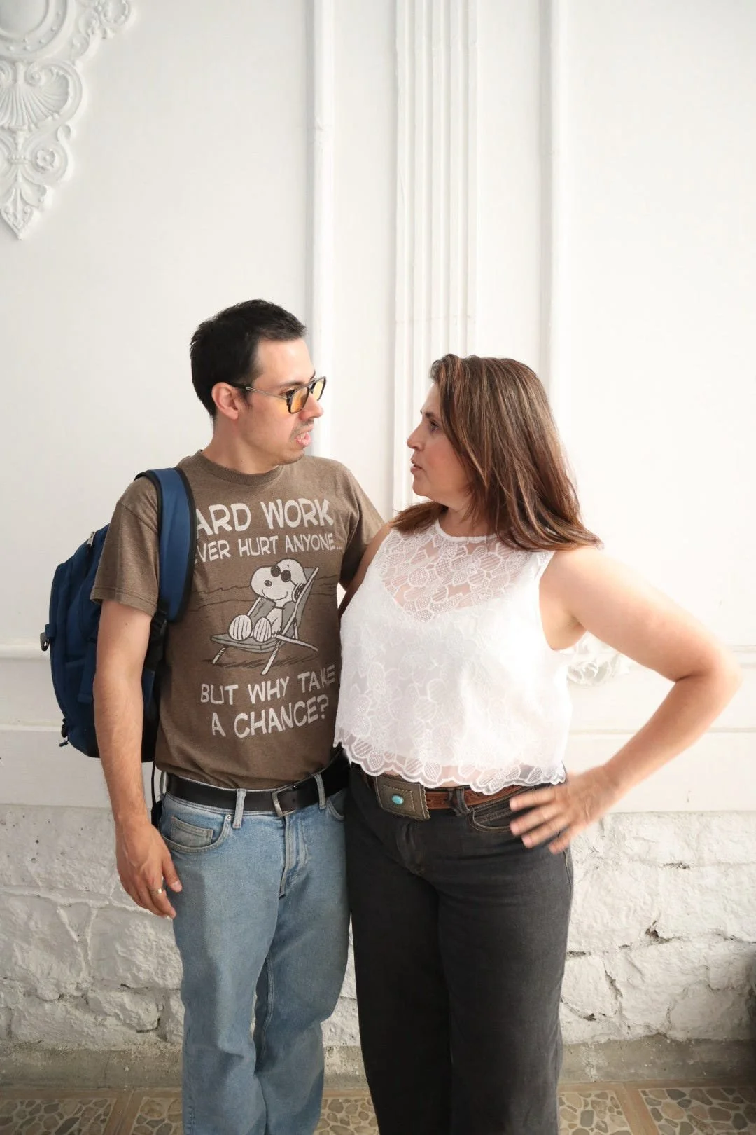 A man and woman stand face-to-face indoors against a white wall with decorative molding, appearing to be in a heated conversation.