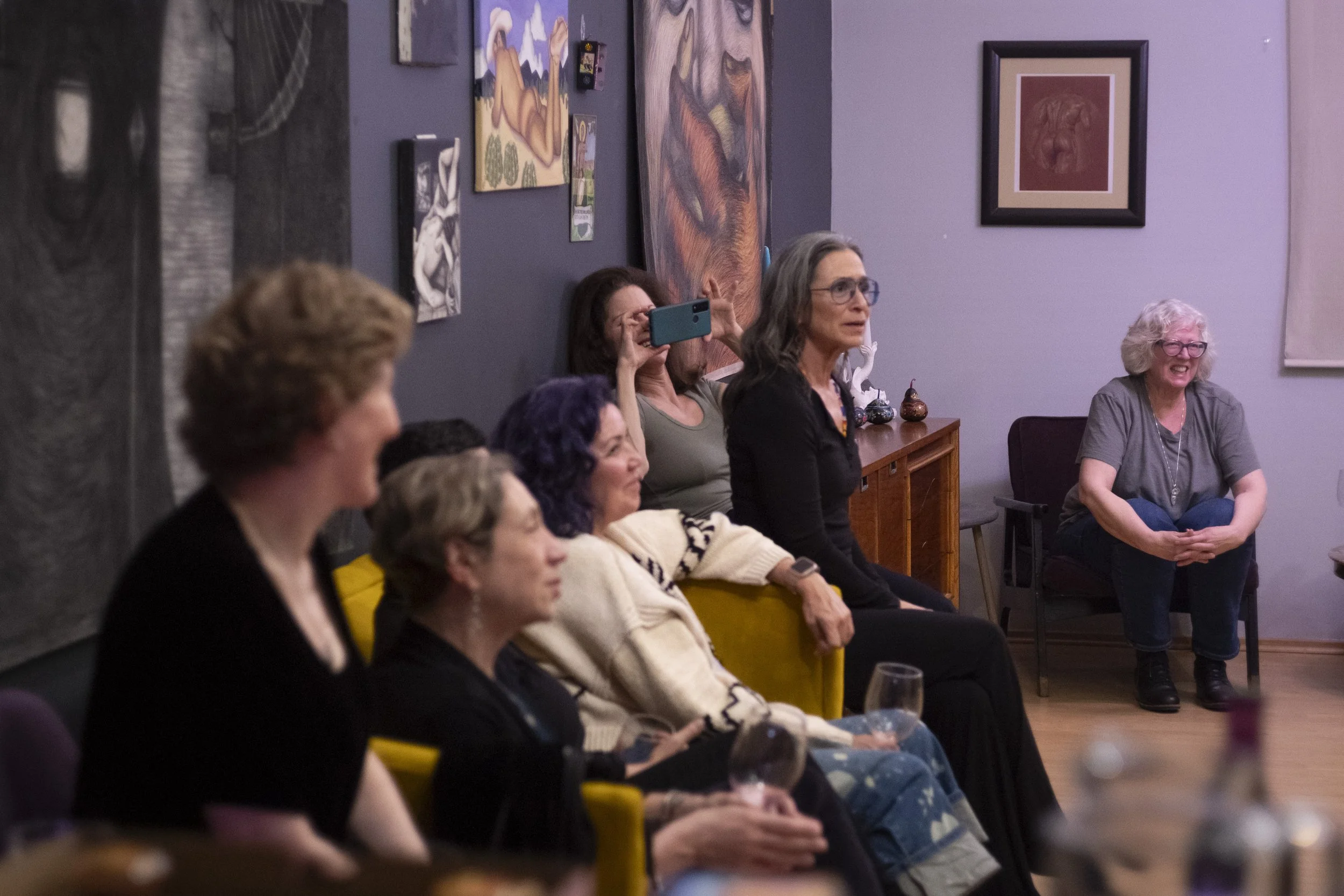 A group of women sitting in chairs in a room with purple walls, some engaging with their phones, others smiling and watching something. The background has various paintings and framed art pieces on the wall.