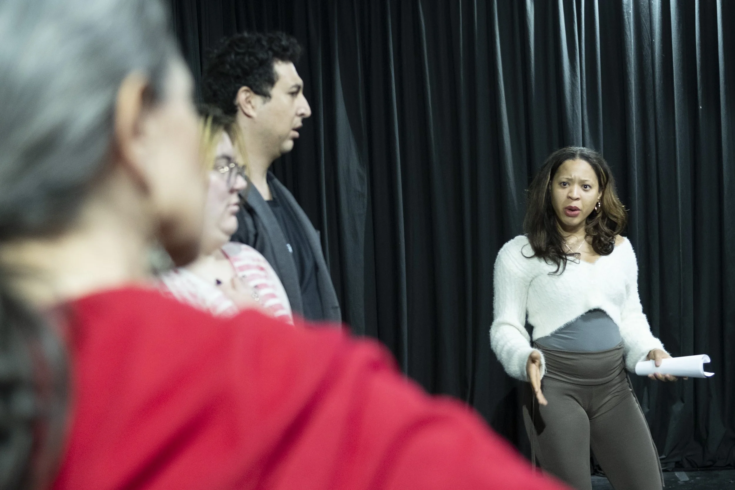 A group of people standing in a room with black curtains, engaged in a discussion or presentation, with a woman in a white sweater and gray skirt holding papers in the foreground. awaken your artist, casa hechale