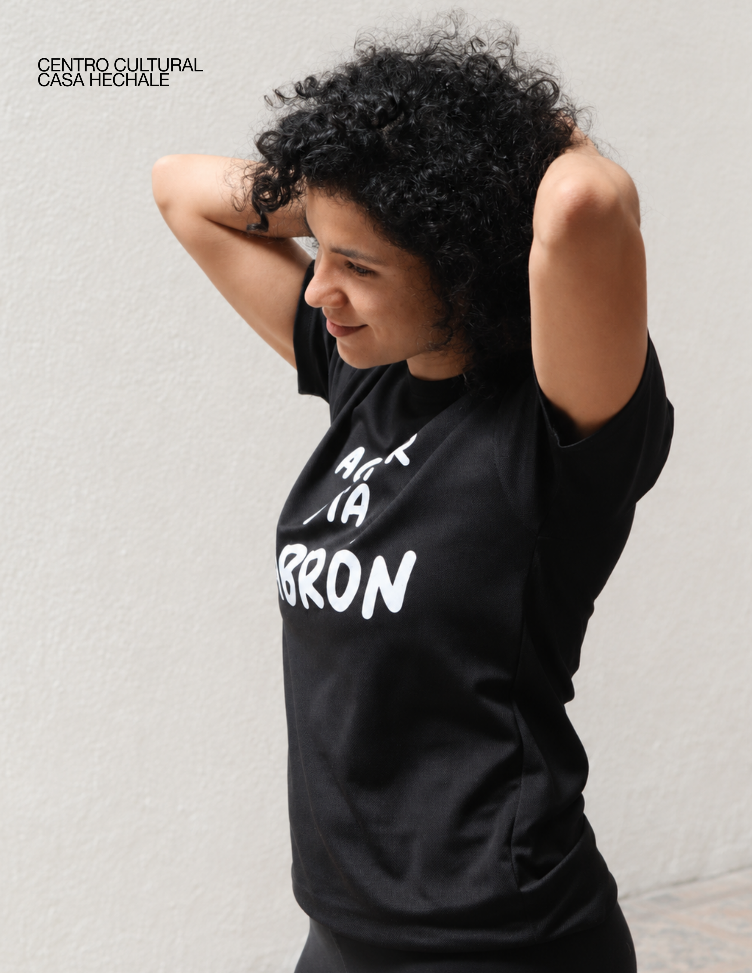 Person with curly dark hair stretching their arms behind their head, wearing a black T-shirt with white text, standing against a plain beige wall. casa hechale