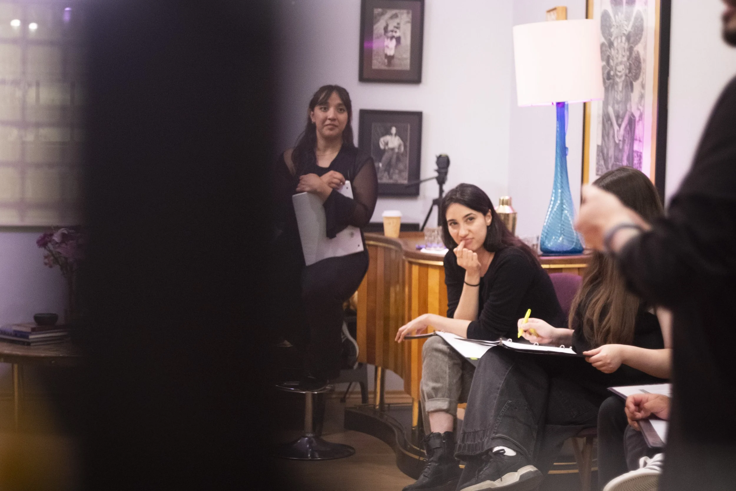 A group of people gathered in a room, with two women sitting and one standing, surrounded by framed artwork on the walls, a tall blue vase, and a lamp. One woman is sitting with her hand on her chin, holding a yellow highlighter, while the standing w