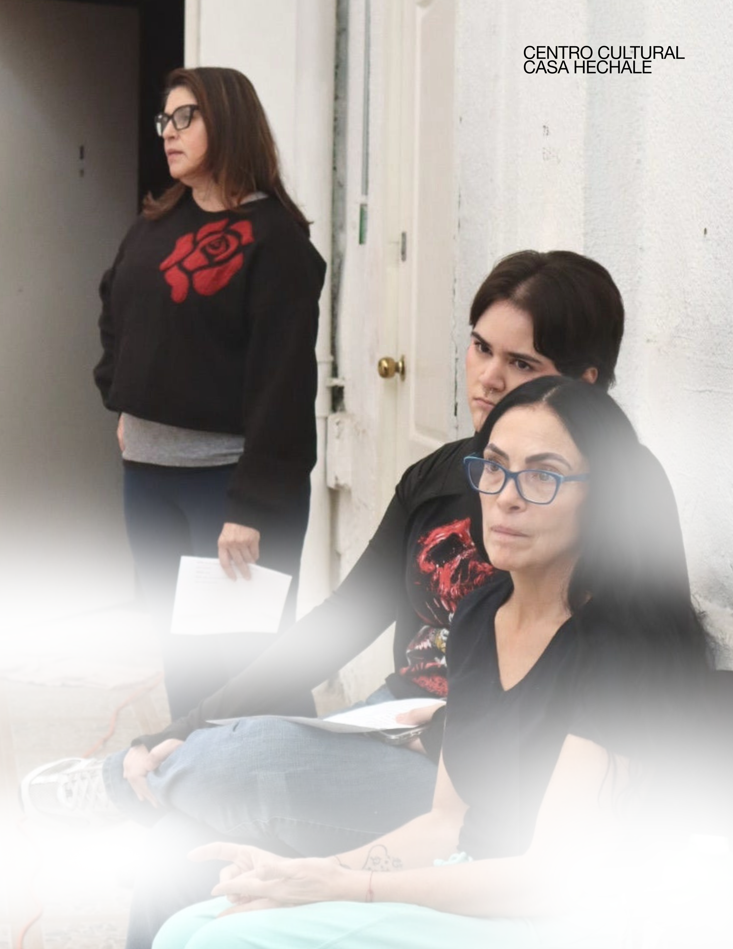 Three women sitting against a white wall with a door, one standing with glasses and a black jacket with a red rose design, two sitting and looking in different directions. awaken your artist, casa hechale