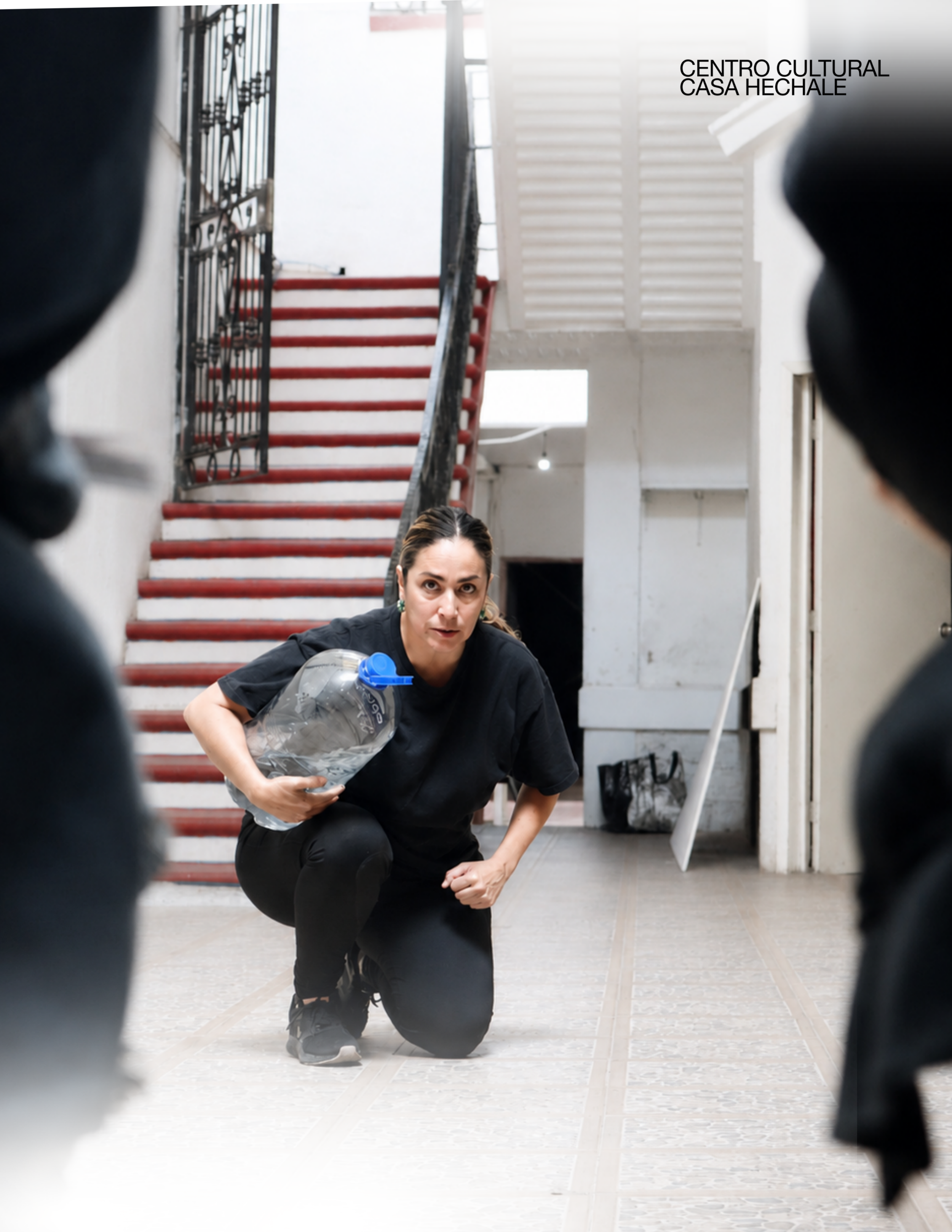 A woman is crouched down and looking directly at the camera, holding a plastic water bottle in her right hand. She is dressed in black and appears to be indoors, with white walls and a red staircase behind her. The setting is identified as the 'Centr