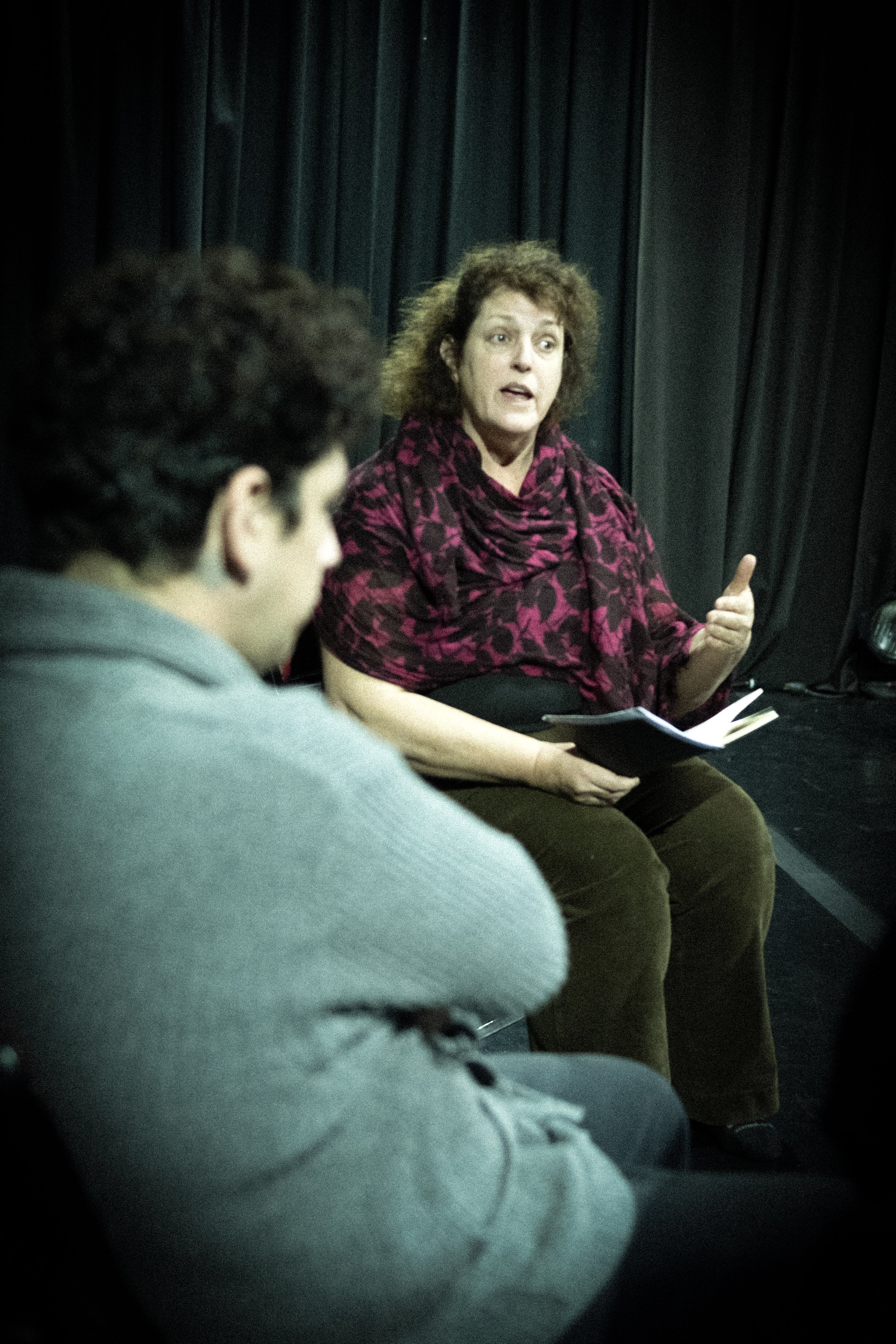 A woman with curly hair wearing a pink and black patterned top is speaking and gesturing with her thumb up, s A person with short dark hair and glasses, dressed in gray, is listening in the foreground. awaken your artist, casa hechale