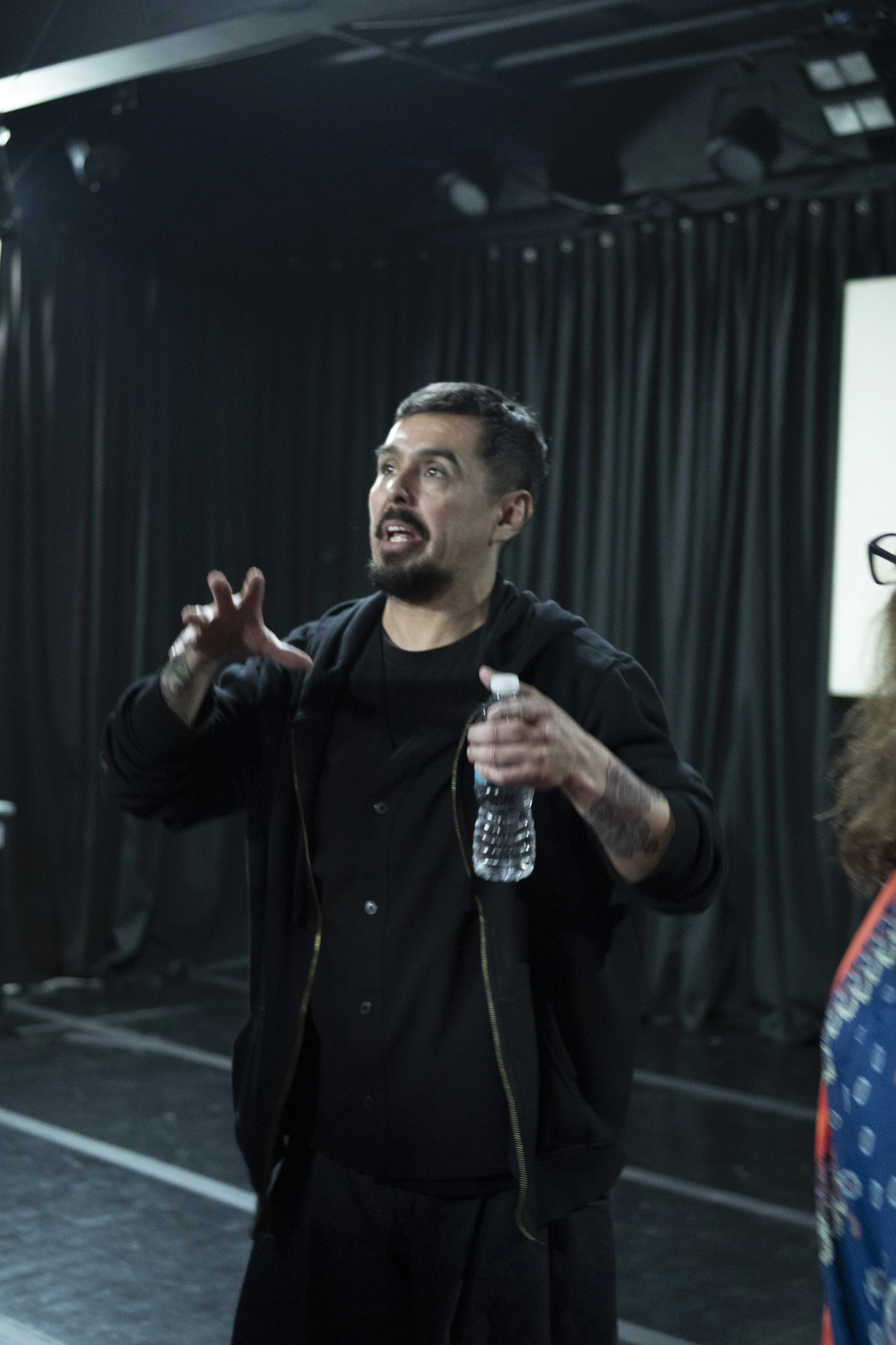 A man with tattoos and a beard speaking, holding a water bottle, in front of dark curtains, possibly at an event or conference. awaken your artist, casa hechale