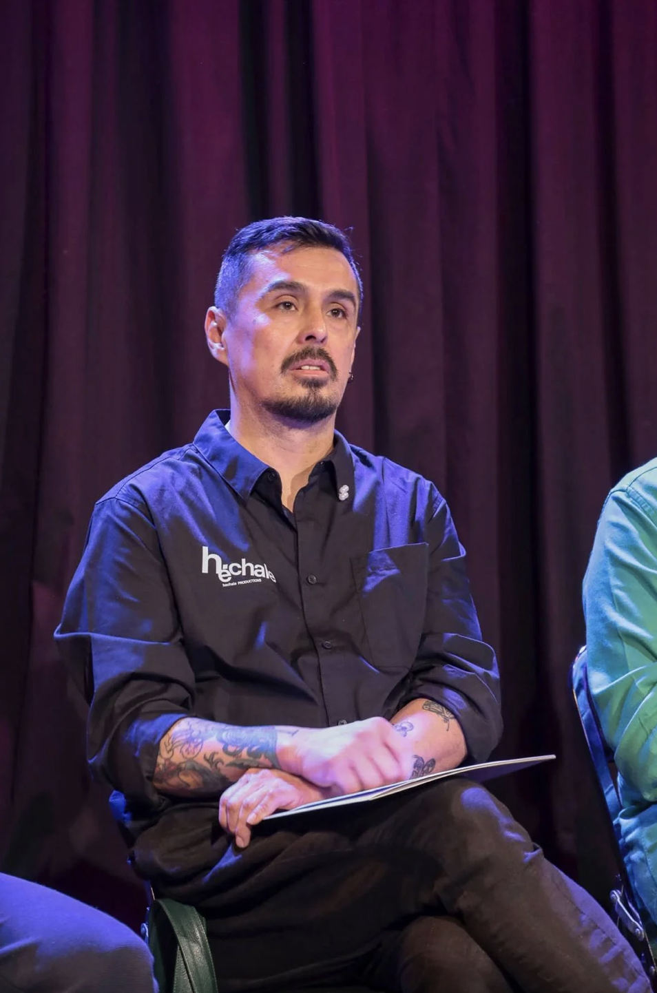 A man with short dark hair, a goatee, and tattoos on his arms, wearing a black shirt with a logo, sitting on a stage in front of dark curtains.