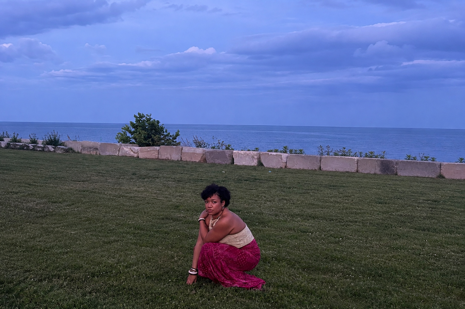 A woman crouching on a grassy area near the ocean, with a stone wall, trees, clouds, and a blue sky in the background.