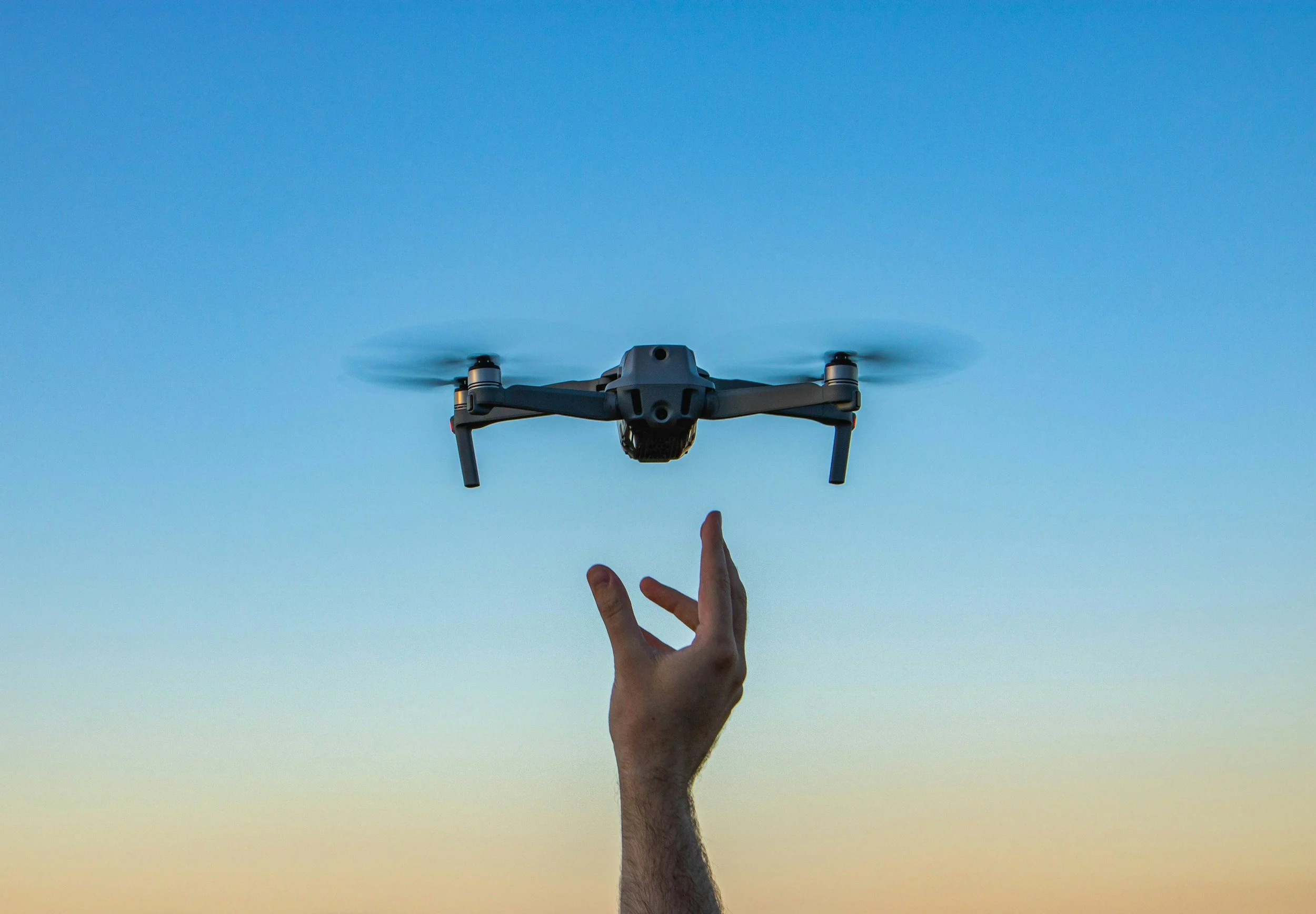 A person holding a drone with their hand, the drone is flying against a clear blue sky.