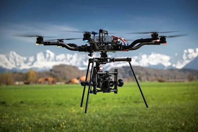 A drone with a camera mounted underneath in an open grass field with mountains in the background.