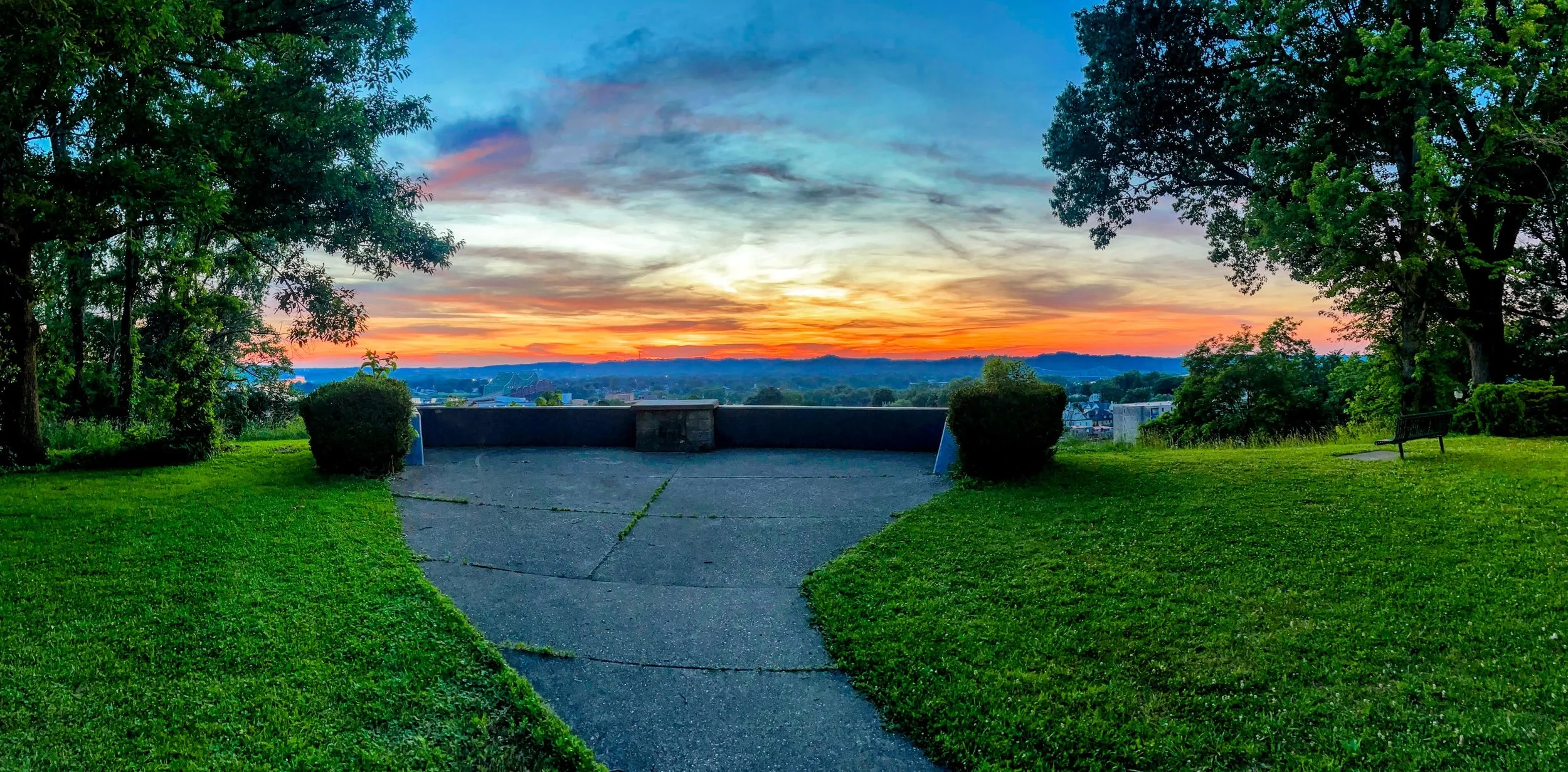 A park at sunset with a paved path, green grass, benches, and trees on either side, overlooking a cityscape and a colorful sky with orange, pink, and blue hues.