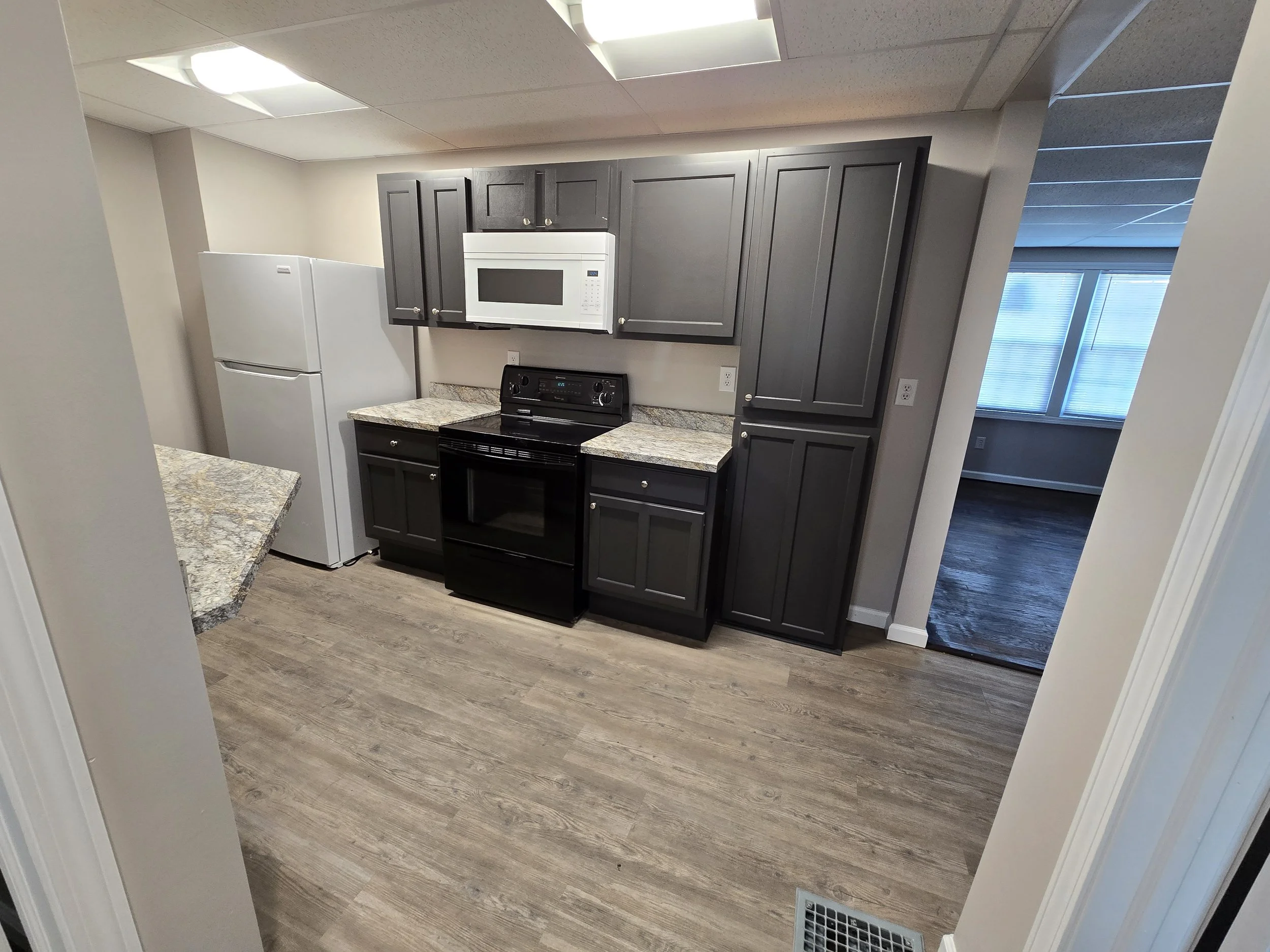 Kitchen with black cabinets, white microwave, black stove, gray countertops, white refrigerator, and windows with blinds.