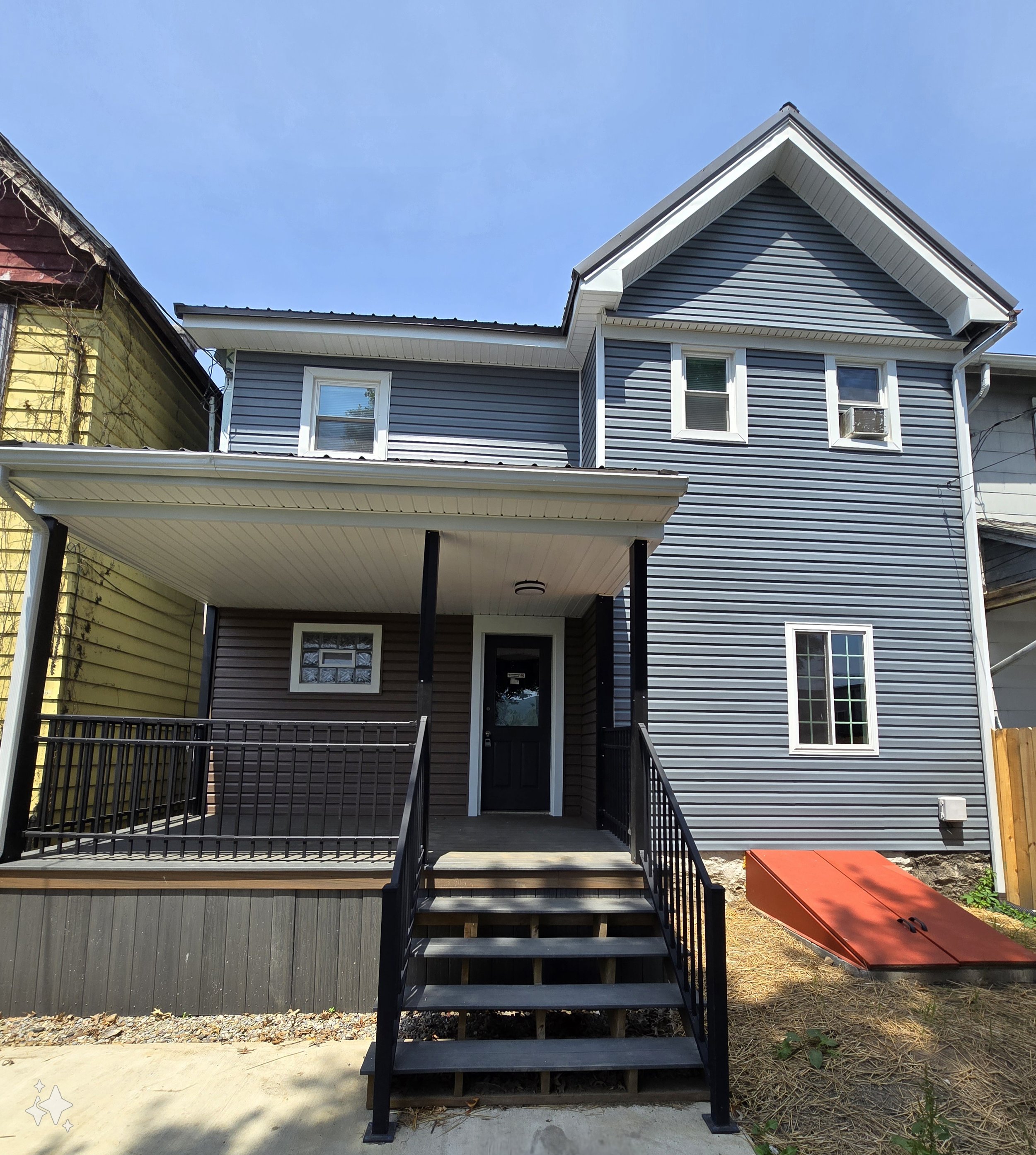 The image shows the back of a house with a blue exterior siding and white trim. There is a small porch with black railings and steps leading up to the door. To the right of the house, there is a red metal object on the ground. The sky above is clear and blue, and the neighboring house with a yellow exterior is partially visible on the left side of the image.