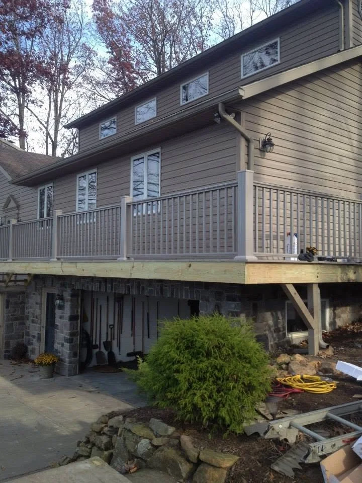 Exterior of a two-story house with a new raised wooden deck, with construction tools and supplies nearby, and trees in the background.