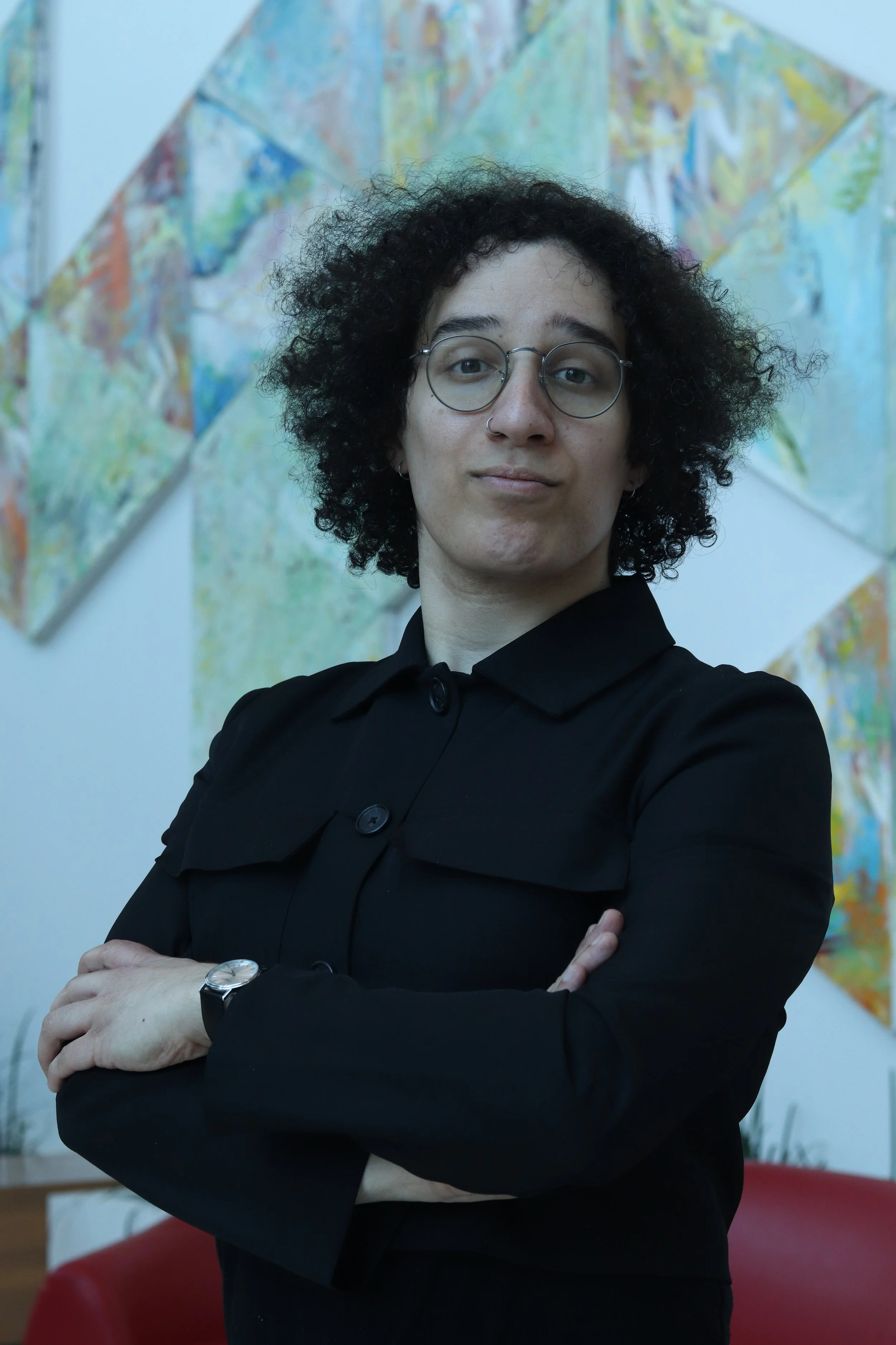 Portrait of a person with curly black hair, glasses, and a nose ring, wearing a black shirt, standing with arms crossed in front of abstract colorful artwork on a white wall.