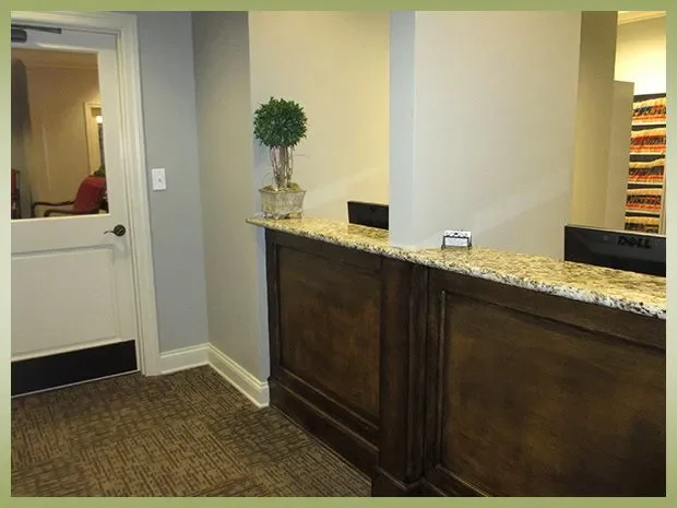 Hotel reception area with a wooden front desk, computer monitors, a potted plant on the counter, and a door leading to a hallway.