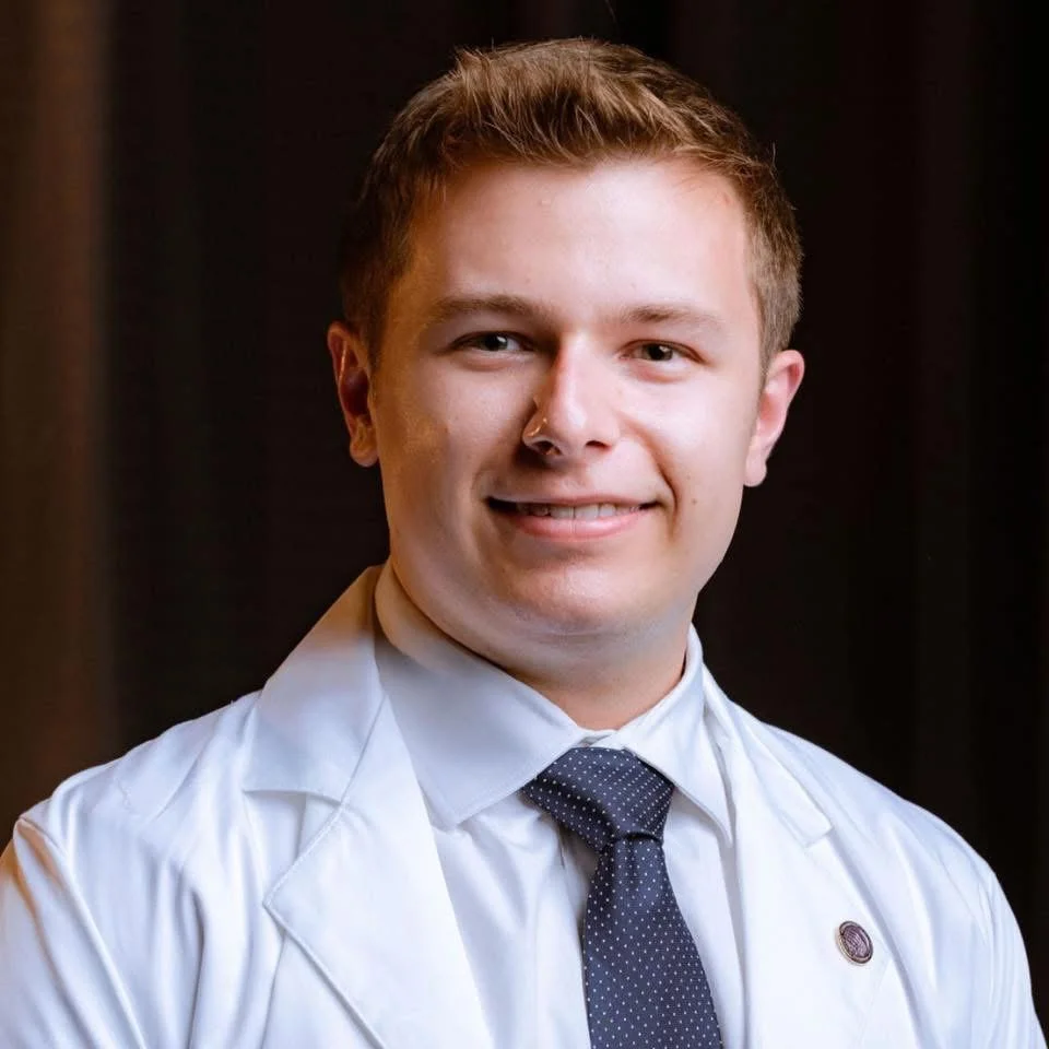Portrait of a young male doctor or medical professional with short light brown hair, wearing a white lab coat, a light-colored shirt, and a dark tie, smiling against a dark background.