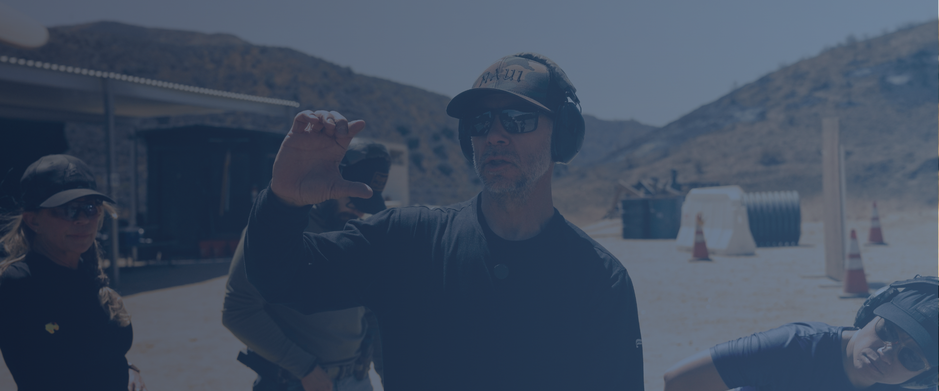 Group of people outdoors in a mountainous area with one man wearing sunglasses, a cap, and headphones, holding his hand up, with other individuals nearby.