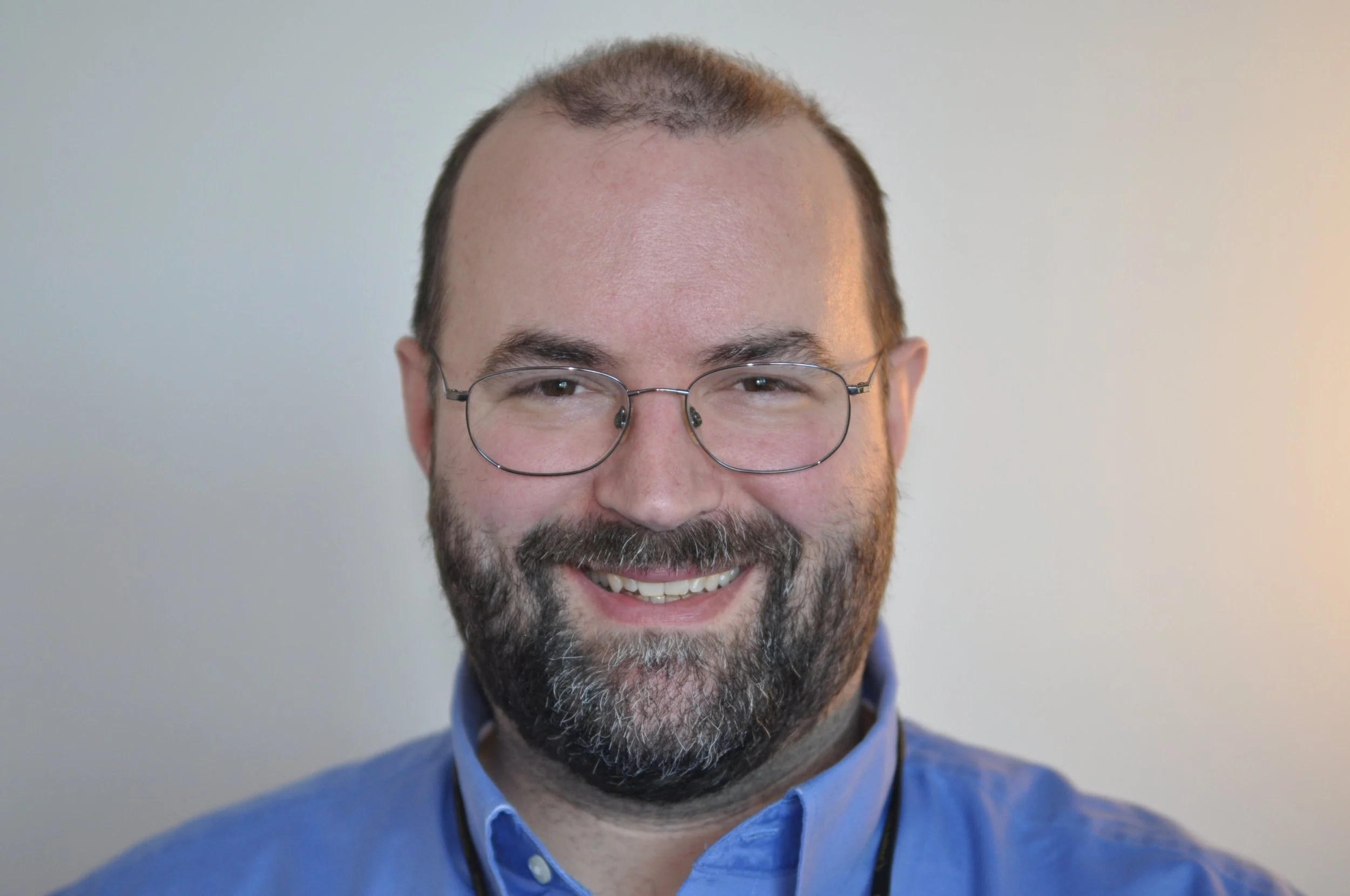 Close-up of a smiling man with glasses, a beard, and short hair, wearing a blue shirt with a white collar. He is indoors with a plain, light-colored background.
