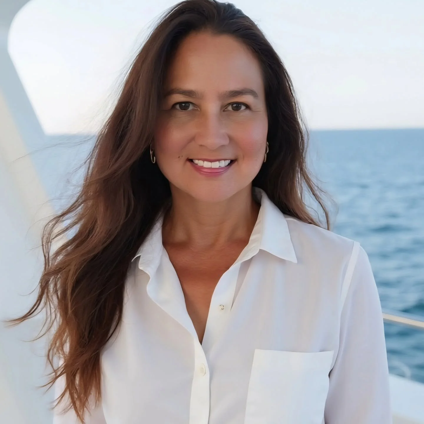 A woman with long, wavy brown hair and tan skin, smiling and wearing a white shirt, standing near the ocean with a blue sky in the background.