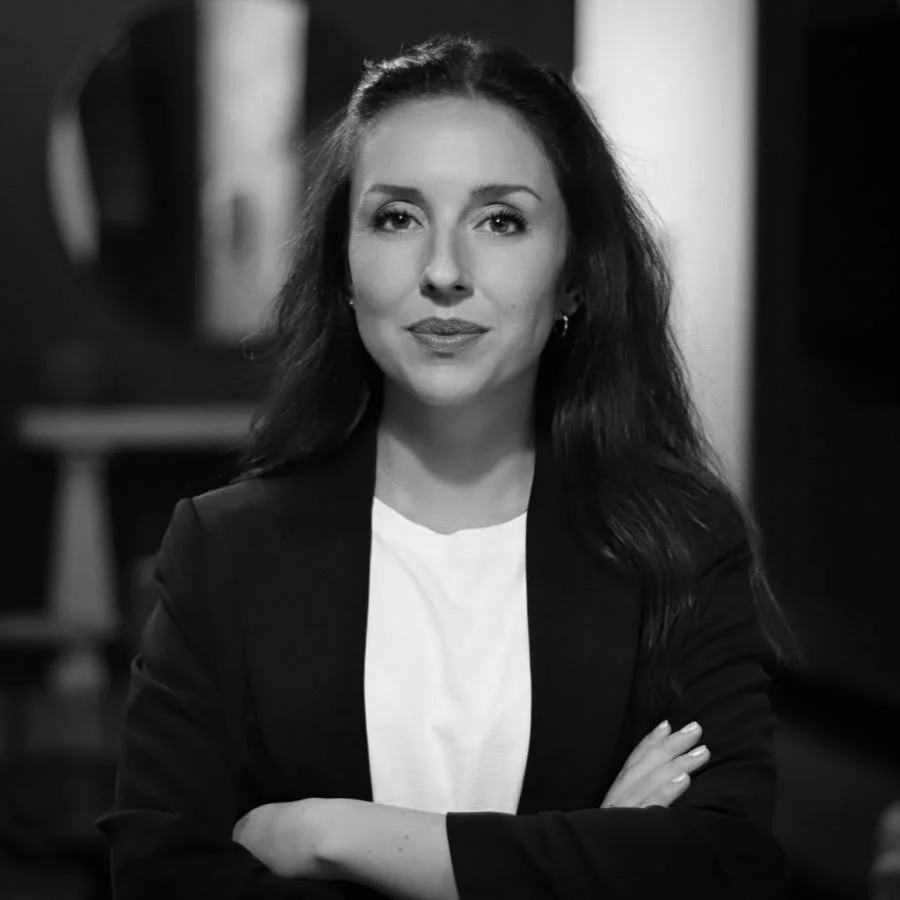 Black and white photo of a confident woman with long wavy dark hair, wearing a blazer and a white shirt, standing indoors with her arms crossed.