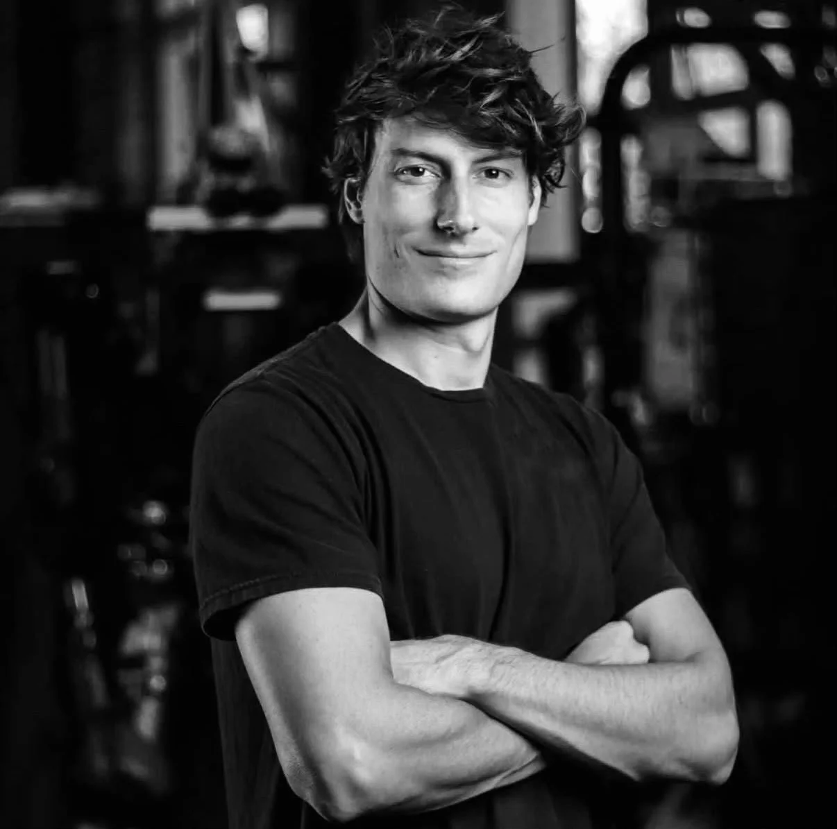 Black and white photo of a young man with wavy hair, smiling, wearing a black t-shirt, standing with arms crossed, in a workshop or similar setting.