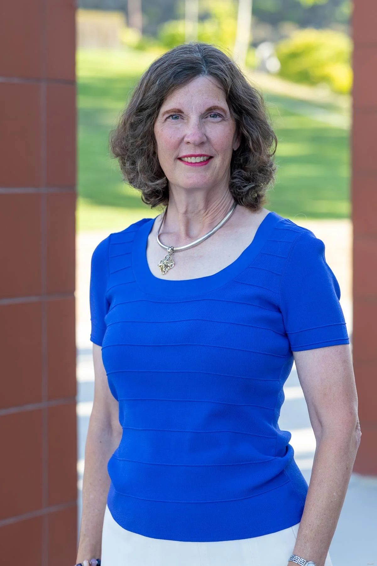 A woman with shoulder-length curly brown hair wearing a bright blue dress and a silver necklace with a pendant, standing outdoors with a blurred green park background.
