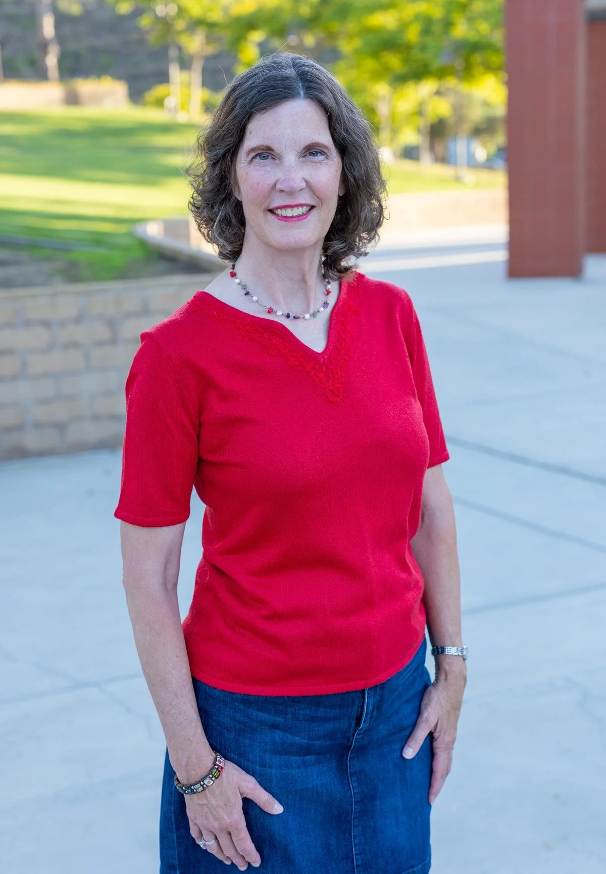 A middle-aged woman with curly brown hair and fair skin smiling outdoors, wearing a red short-sleeved top, blue jeans, and colorful jewelry, with trees and a brick structure in the background.