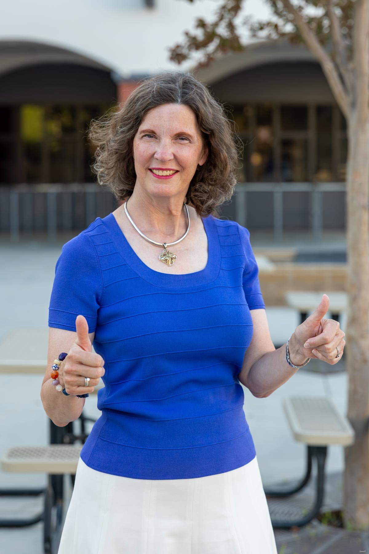 A woman with curly gray hair, wearing a blue dress and a silver necklace, giving two thumbs up outdoors, with trees and picnic tables in the background.