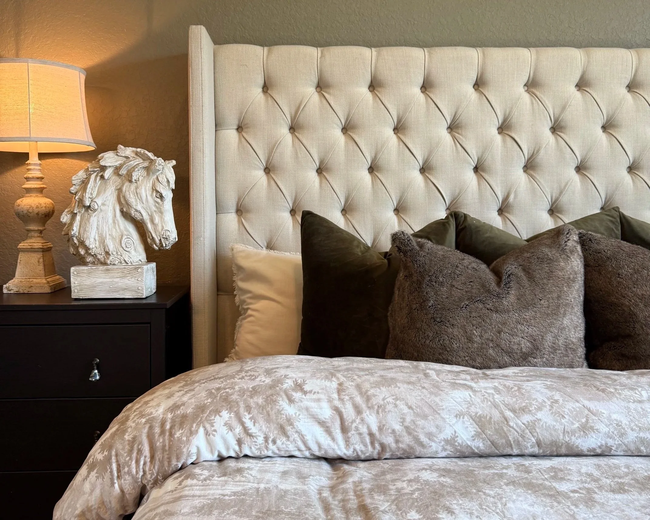 A bedroom with a beige tufted headboard, brown and beige pillows, a nightstand with a sculpted animal head lamp, and a beige bedspread.