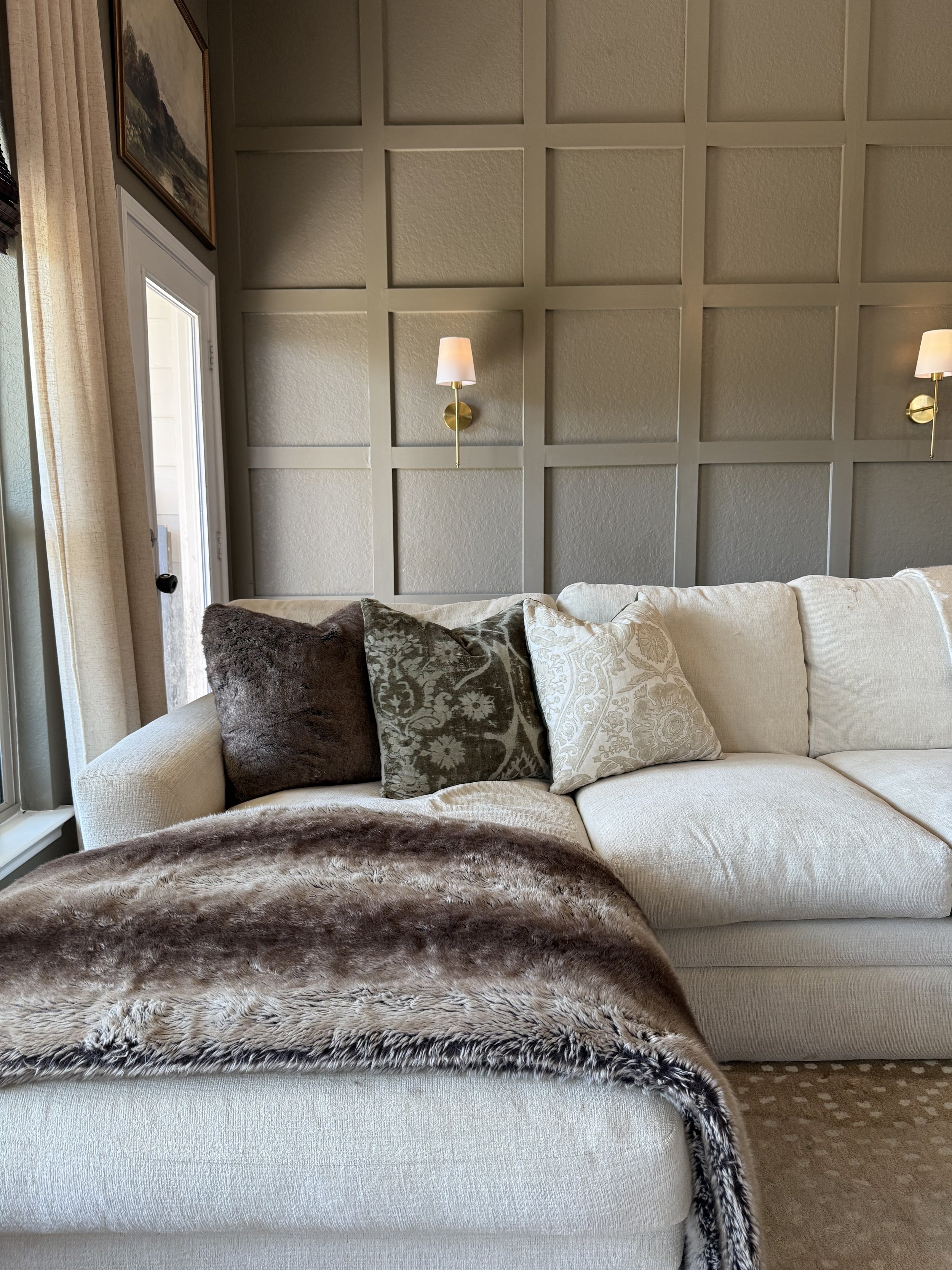 Living room with a cream-colored sofa, decorative pillows, a faux fur throw, wall sconces, a window with curtains, and a wall with square paneling.