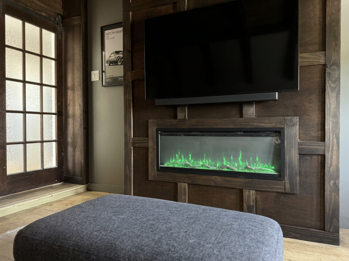 A living room corner with a mounted flat-screen TV above an electric fireplace with green flames, a gray ottoman in the foreground, a glass door to the left, and a framed car poster on the wall.