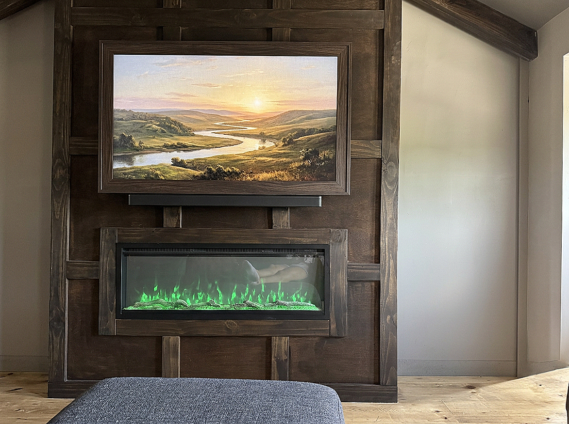 A living room features a wooden wall with a framed landscape painting of a river and sunset, an electric fireplace with green flames, and a gray upholstered sofa in the foreground.