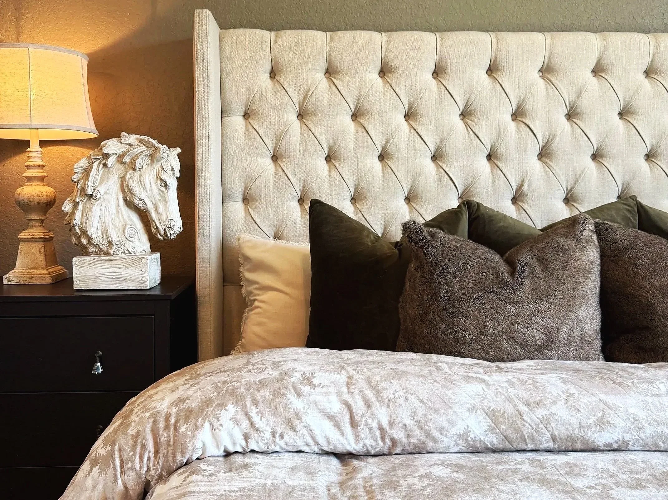 A cozy bedroom scene featuring a large tufted beige headboard, a bedside table with a lamp, a sculpture of a lion's head, and a bed with a cream-colored patterned duvet and assorted pillows.