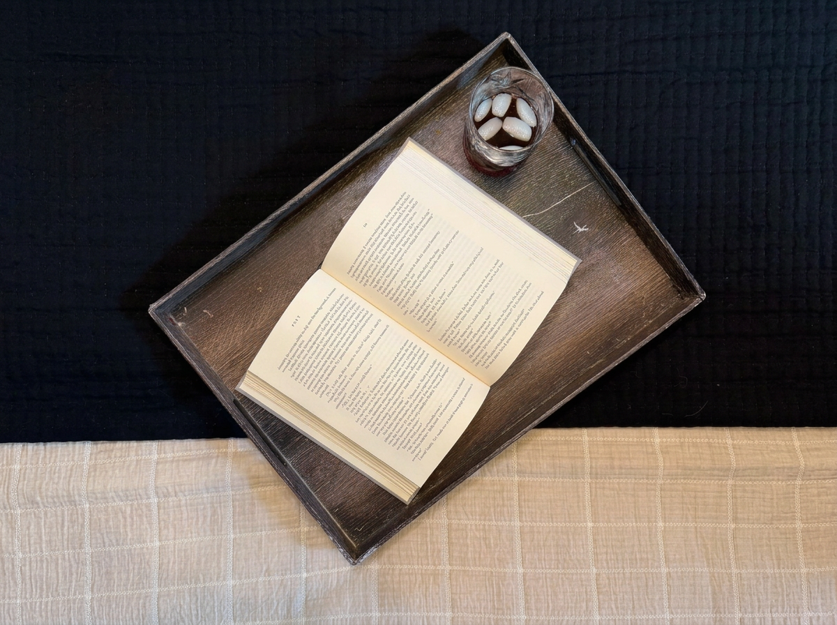 An open book on a tray, next to a glass of water with ice cubes, on a surface with a black and beige tablecloth.