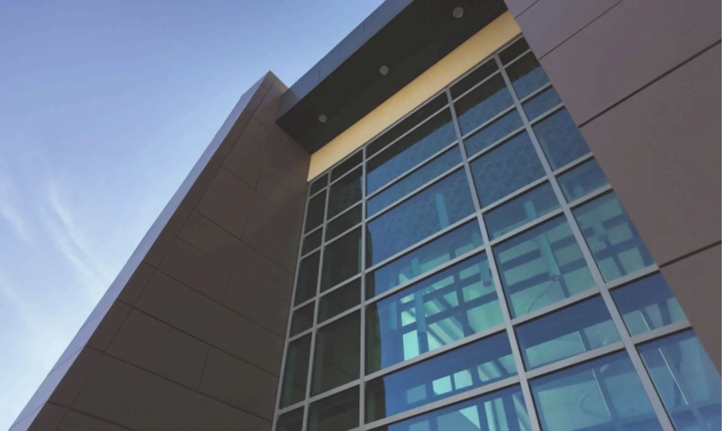 Low-angle view of a modern building with large glass windows and beige exterior panels, under a clear blue sky.
