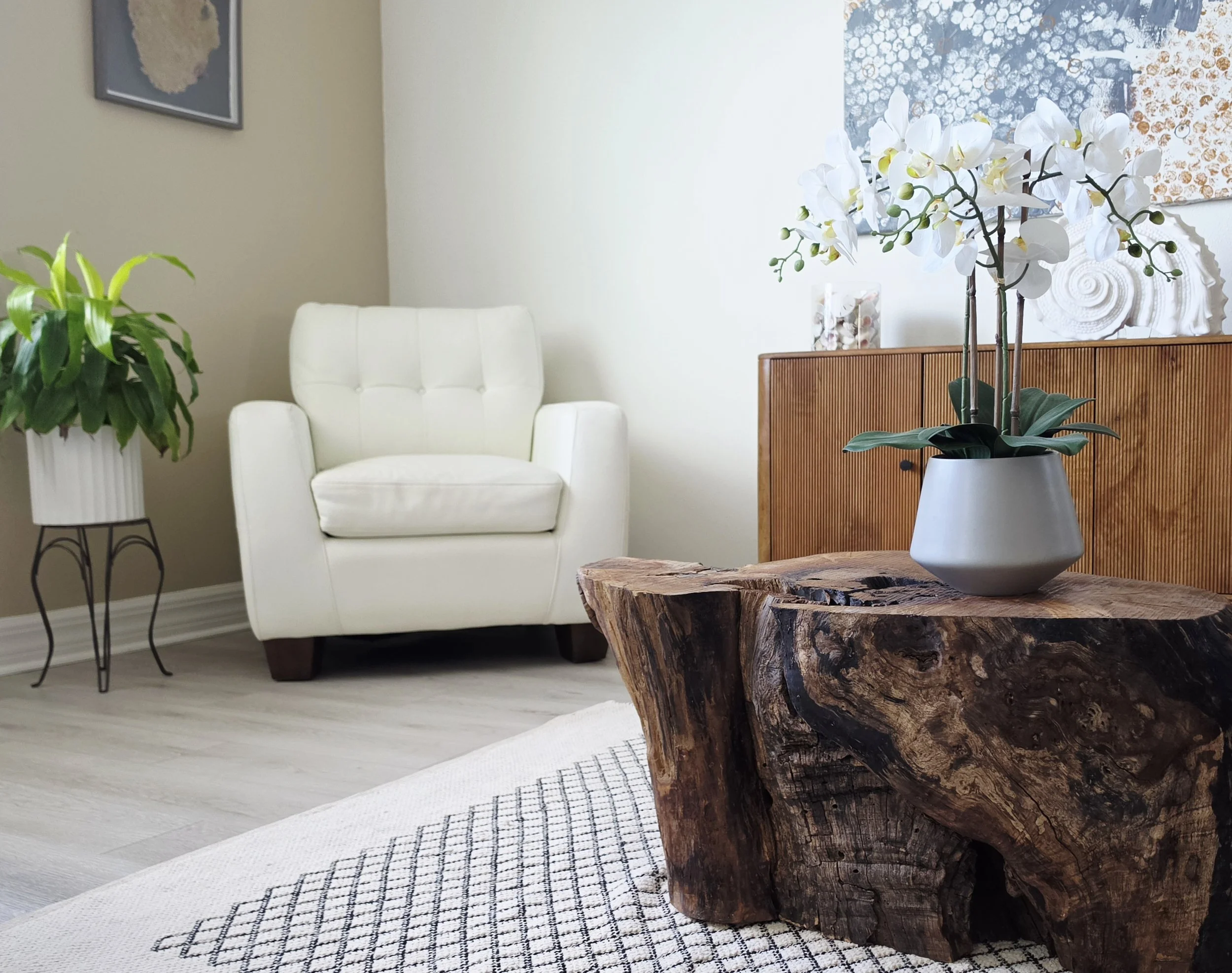 Interior living space with a white armchair, a small table with a large potted orchid on it, a decorative wooden sideboard, two pieces of wall art, and some additional houseplants.