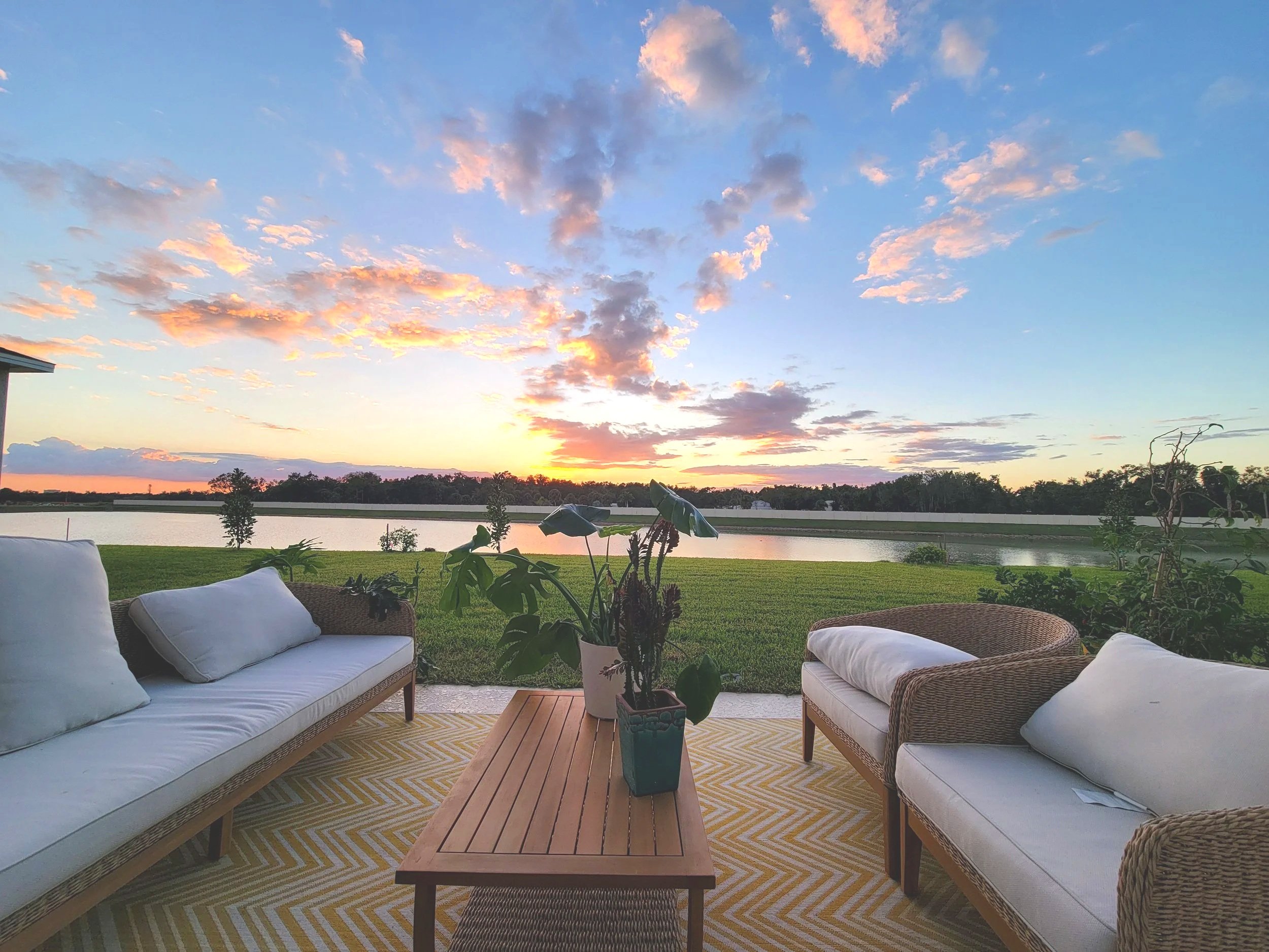 An outdoor patio area with white cushioned chairs and wicker sofas, a wooden coffee table, and potted plants, overlooking a serene lake with a sunset sky filled with colorful clouds.