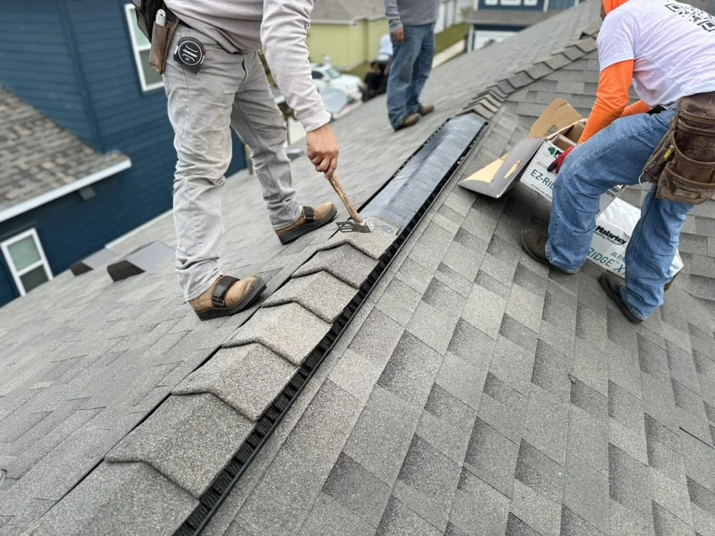 Roofers installing shingles and a snow guard on a residential roof.