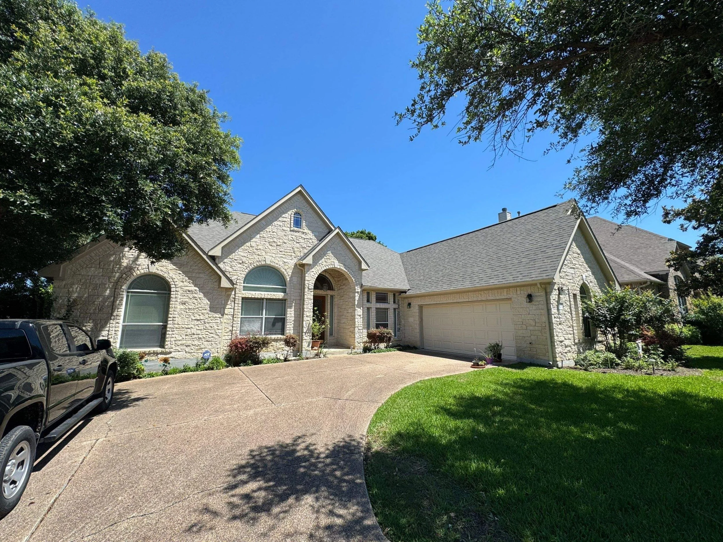 A single-story house with a stone exterior, arched windows, and a driveway in front, surrounded by greenery and trees, under a clear blue sky.