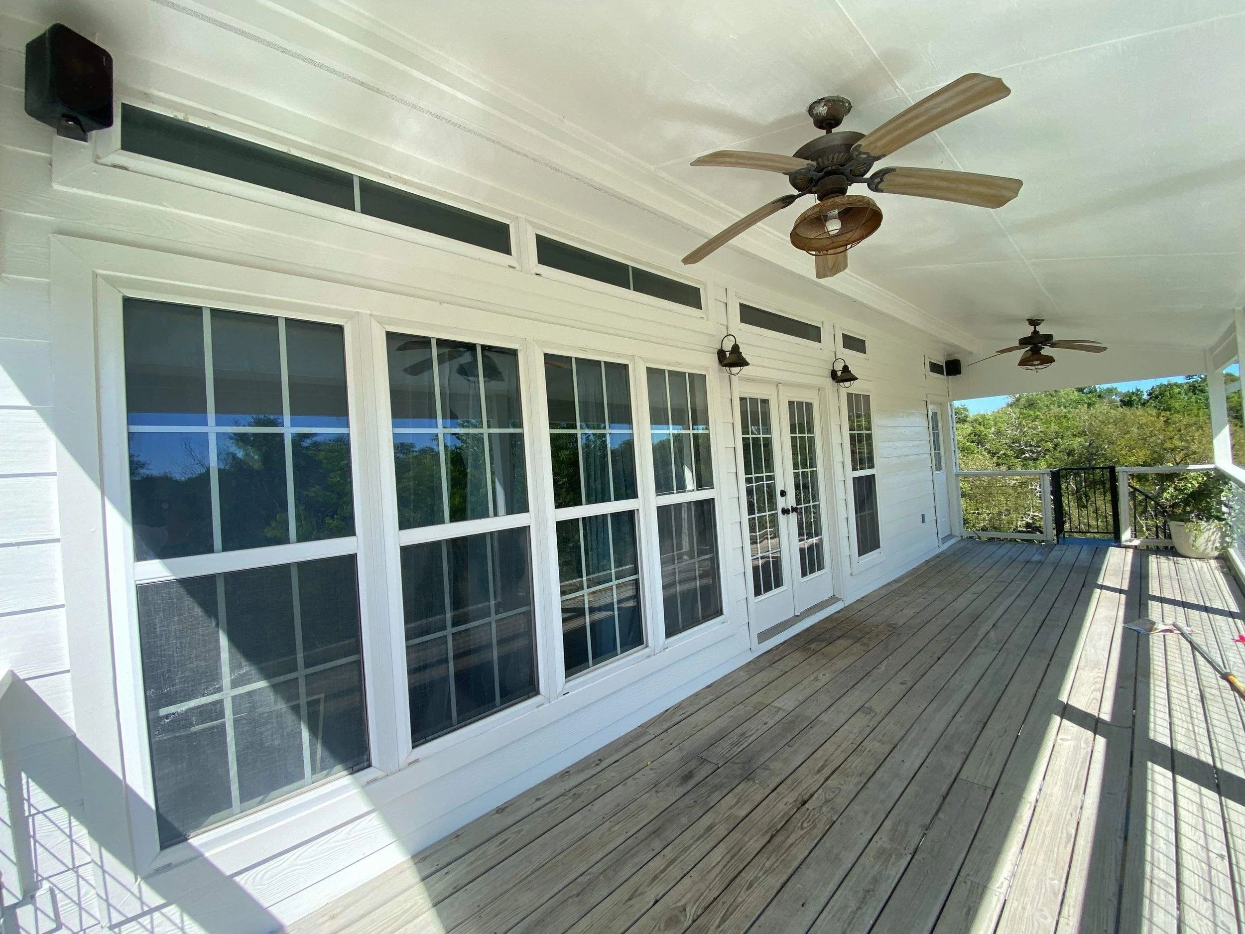 Empty outdoor balcony with wooden flooring, white siding walls, multiple windows, two ceiling fans, and a potted plant in the corner, with greenery visible beyond the railing.