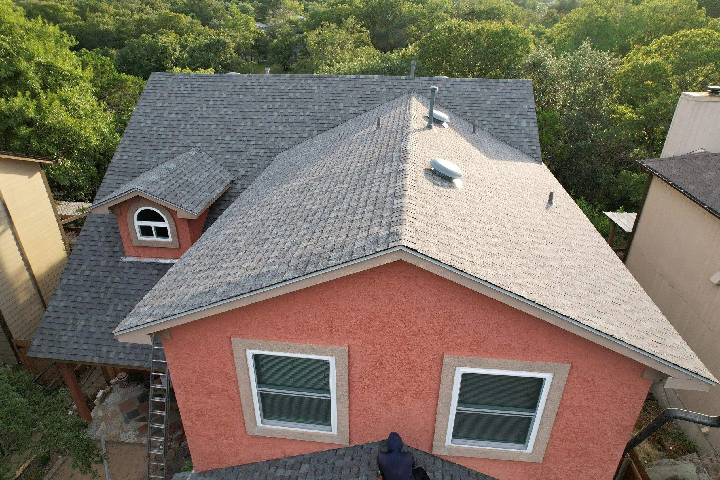 View from above of a house with a pink exterior, gray shingle roof, and two double-pane windows. Surrounding trees are visible in the background.