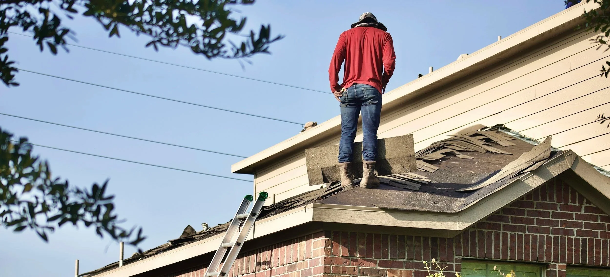 A person standing on a ladder on the roof of a brick house, working on the shingles, with trees and a blue sky in the background.