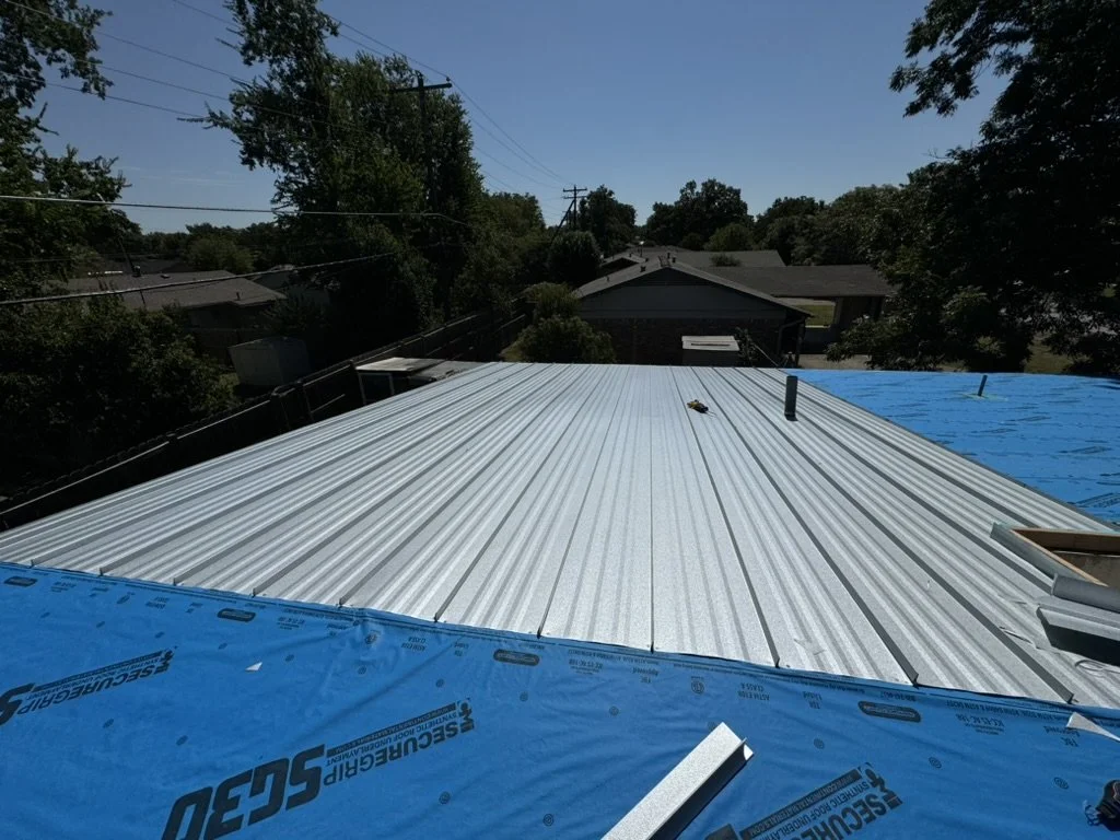 Roof with metal panels installed, some blue roofing underlayment visible, surrounded by trees and neighboring houses.
