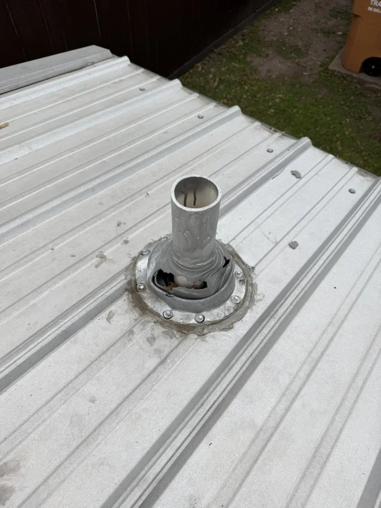 Metal vent pipe protruding from the roof of a building with a weathered metal surface, surrounded by a circular metal flange with bolts.
