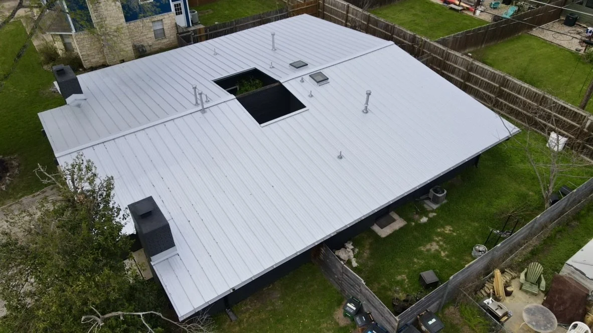 Aerial view of a house with a shiny metal roof, multiple vents, skylights, and a cutout section, surrounded by a wooden fence, with neighboring houses and green lawns.
