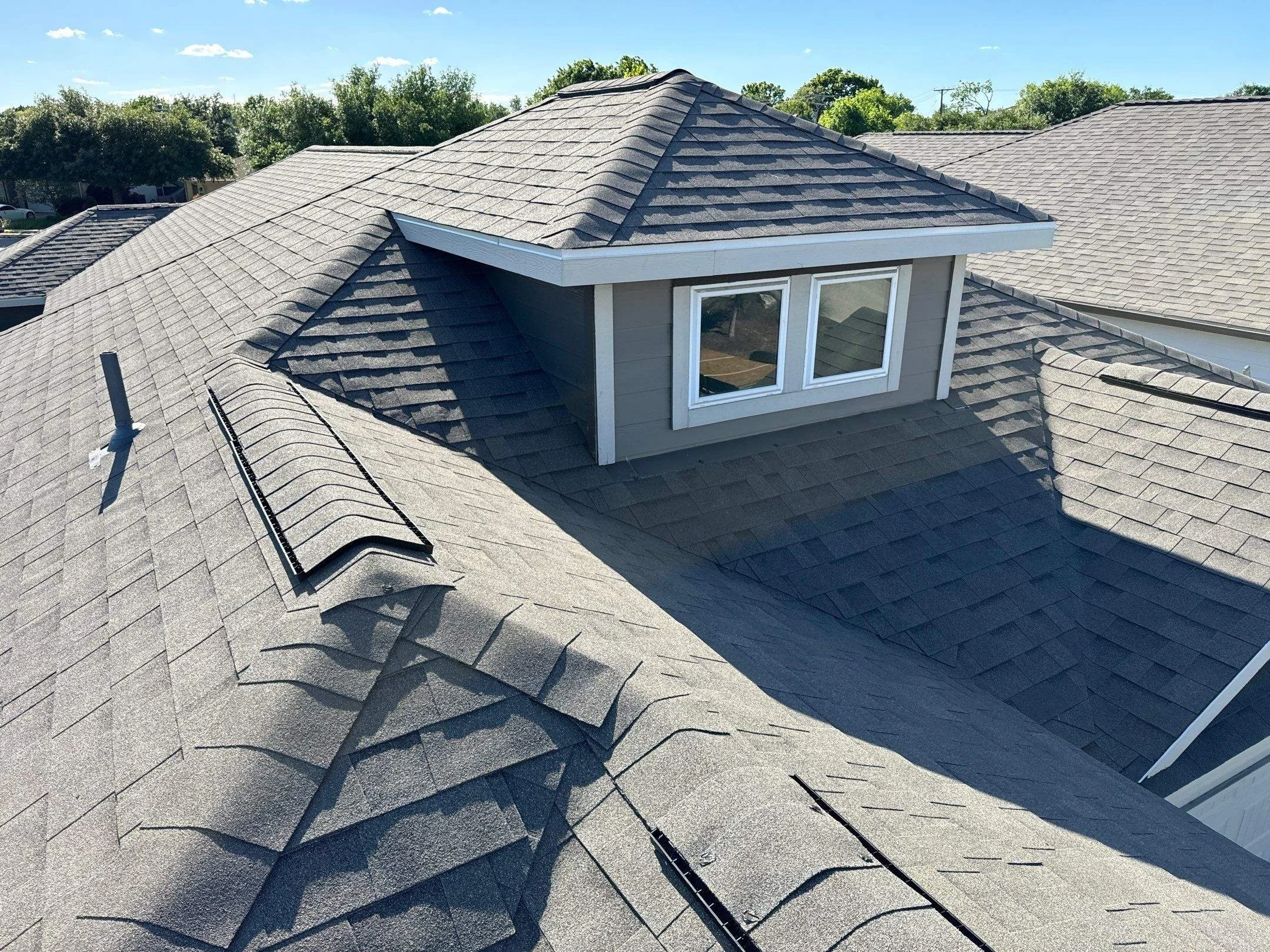 Aerial view of a house roof with multiple gables and dormer window, covered in gray asphalt shingles, with trees in the background and a clear blue sky.