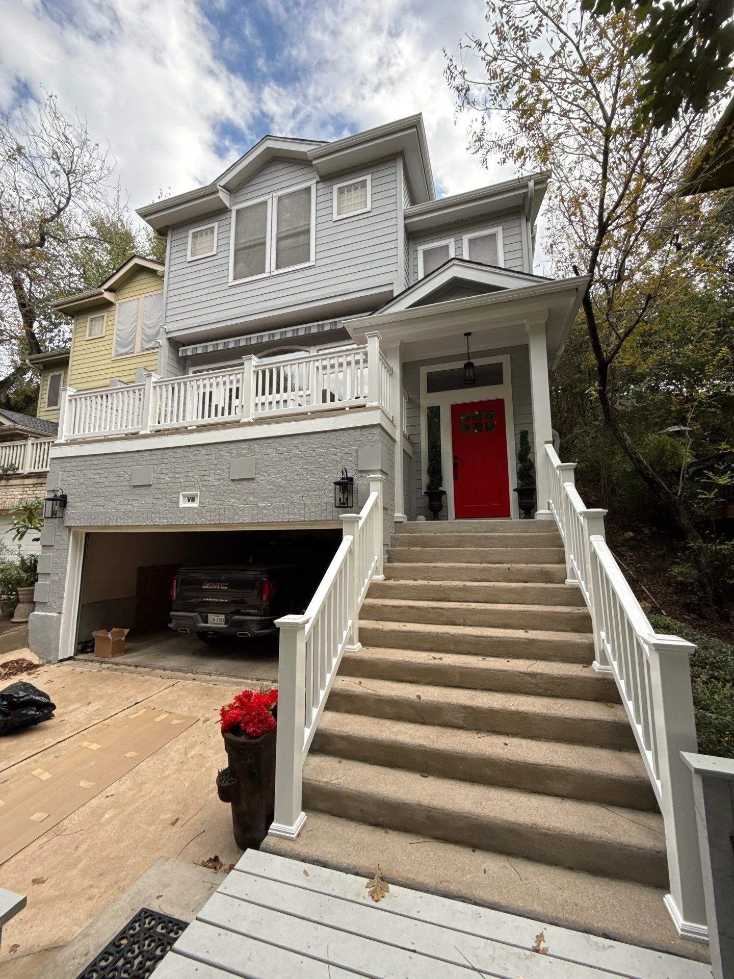 Multi-story house with gray and yellow siding, red front door, and white railing stairs leading up to the entrance. A garage with a GMC truck and a black trash bag in front. Potted red flowers are on the porch. Trees surround the house under a partly cloudy sky.